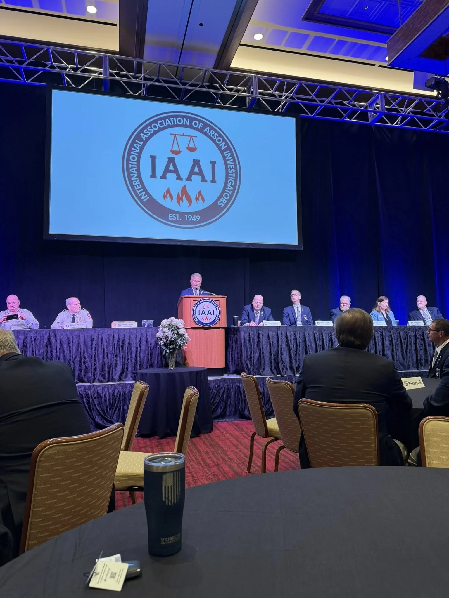 A large conference room with a stage at the front, where a panel of people is seated behind a long table draped with purple cloth. There is a speaker at a podium with the IAAI logo on it, speaking into a microphone. A large screen above displays the 