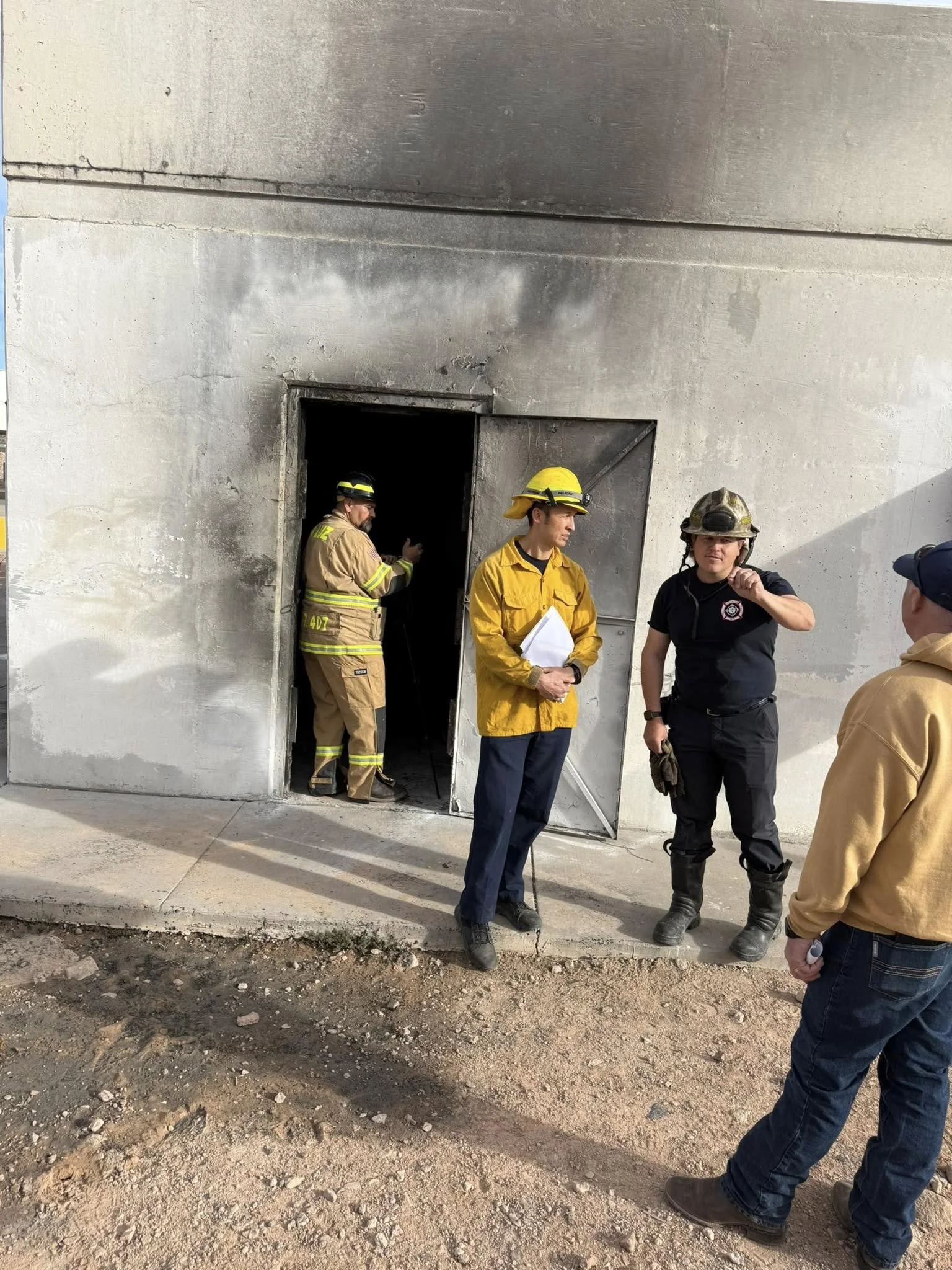 Group of firefighters and emergency personnel standing outside a building with a burned or damaged door. They are engaged in a discussion, some taking notes or inspecting the scene.