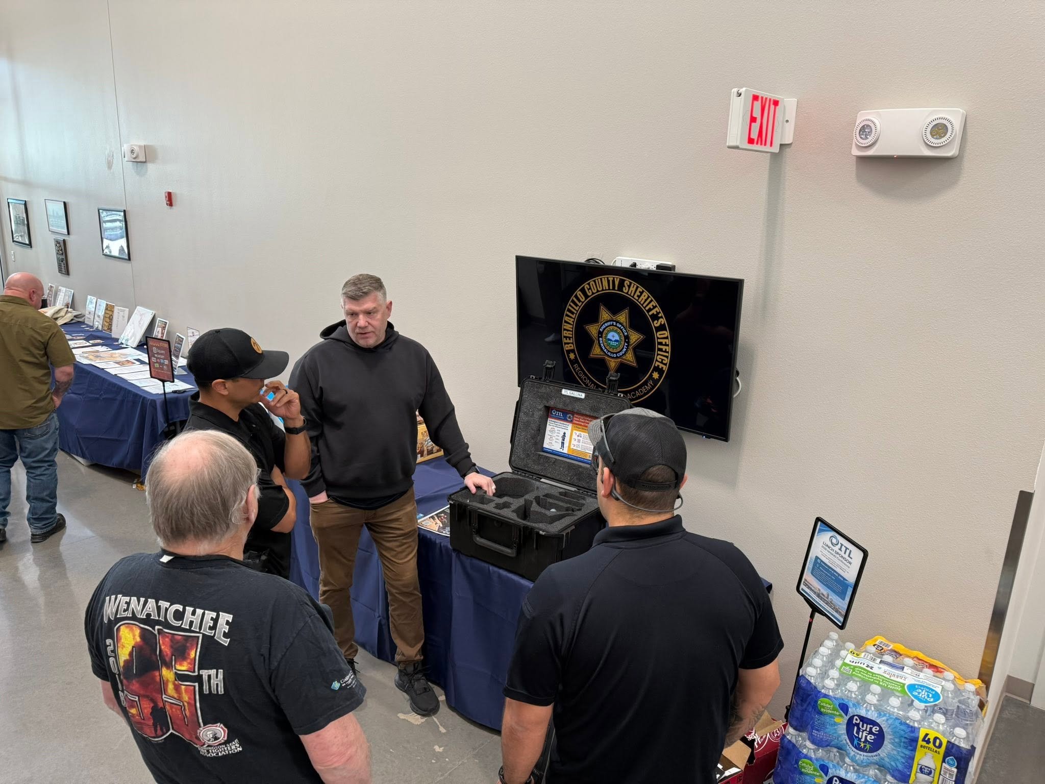 A group of people gathered around a display with the Beverly Willo County Sheriff's Office logo, some looking at items and engaging in conversation, in an indoor setting with a table of pamphlets and informational materials.