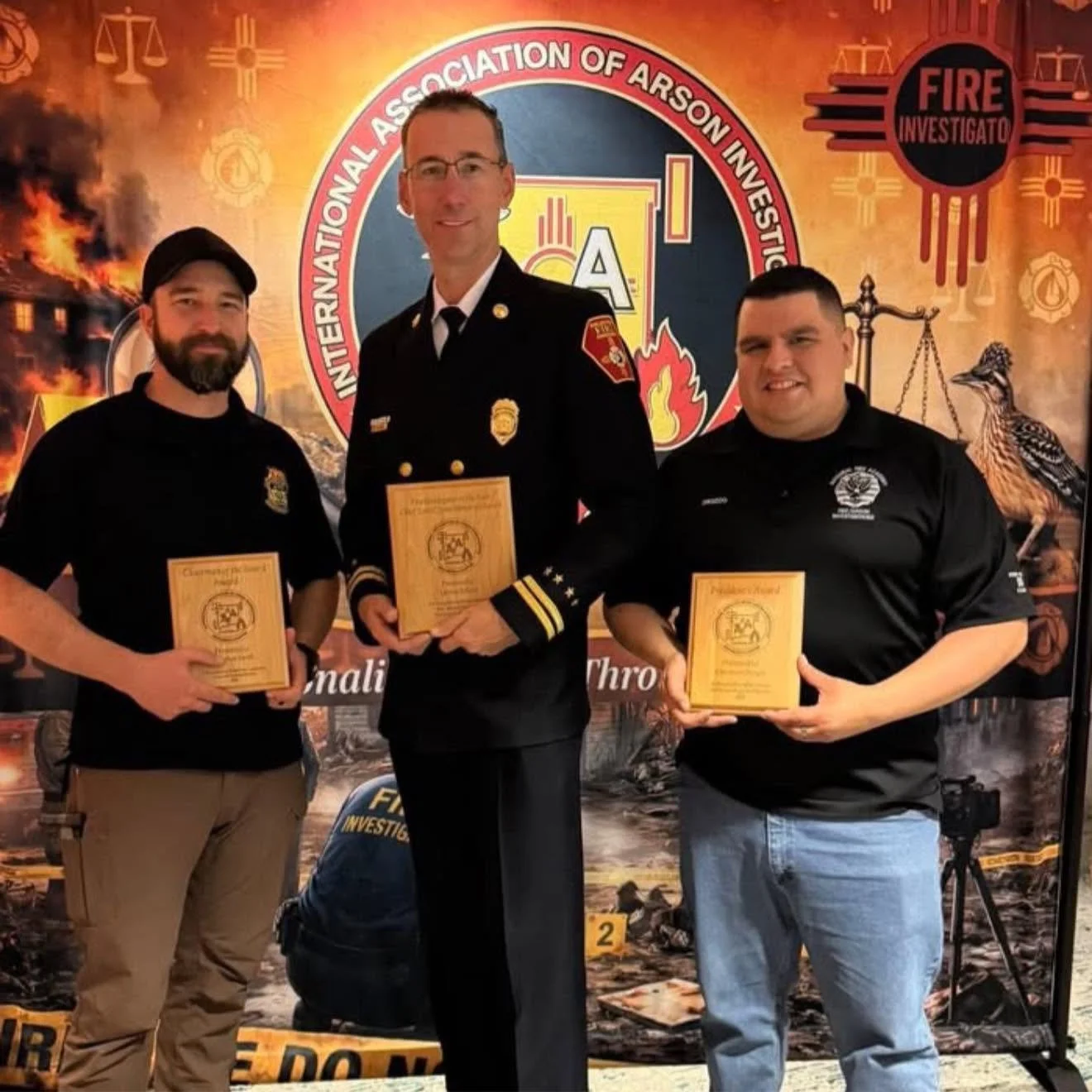 Three men standing together in front of a backdrop with the logo of the International Association of Arson Investigators, holding plaques. The central man is in a fire department uniform. The backdrop features firefighting and law enforcement symbols