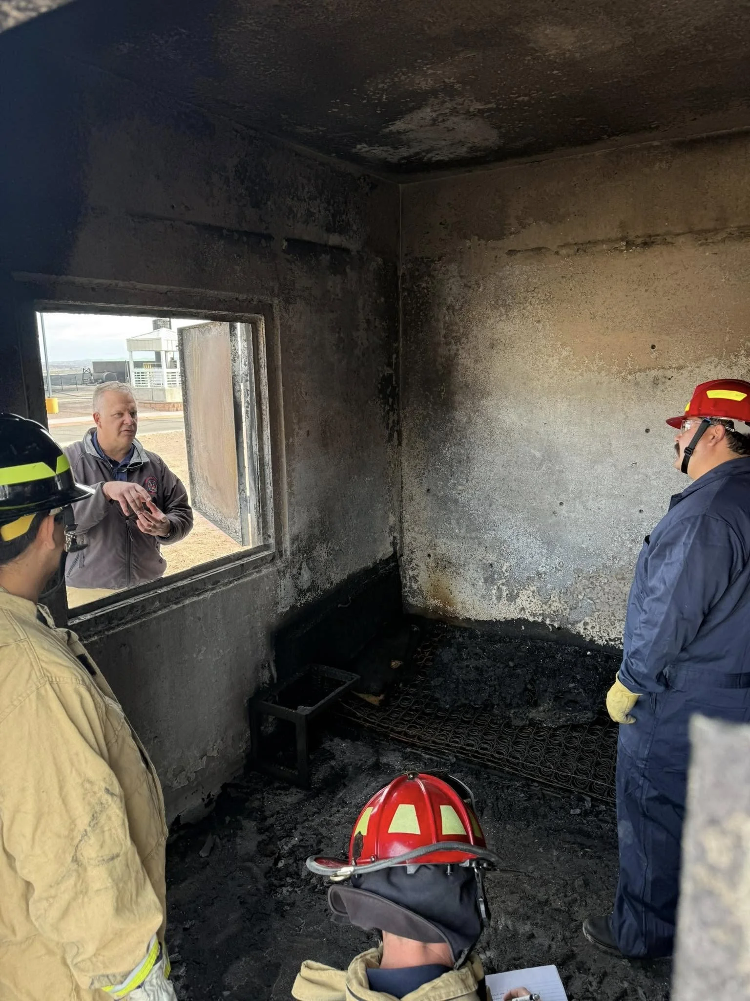 Firefighters and a man in a burned, charred room with blackened walls and a window, some firefighters wearing helmets and protective gear.