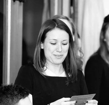 Black and white photo of Katrin Winstanley, wedding celebrant with shoulder-length hair smiling as she reads vows to the couple getting married. With other people partially visible in the background.