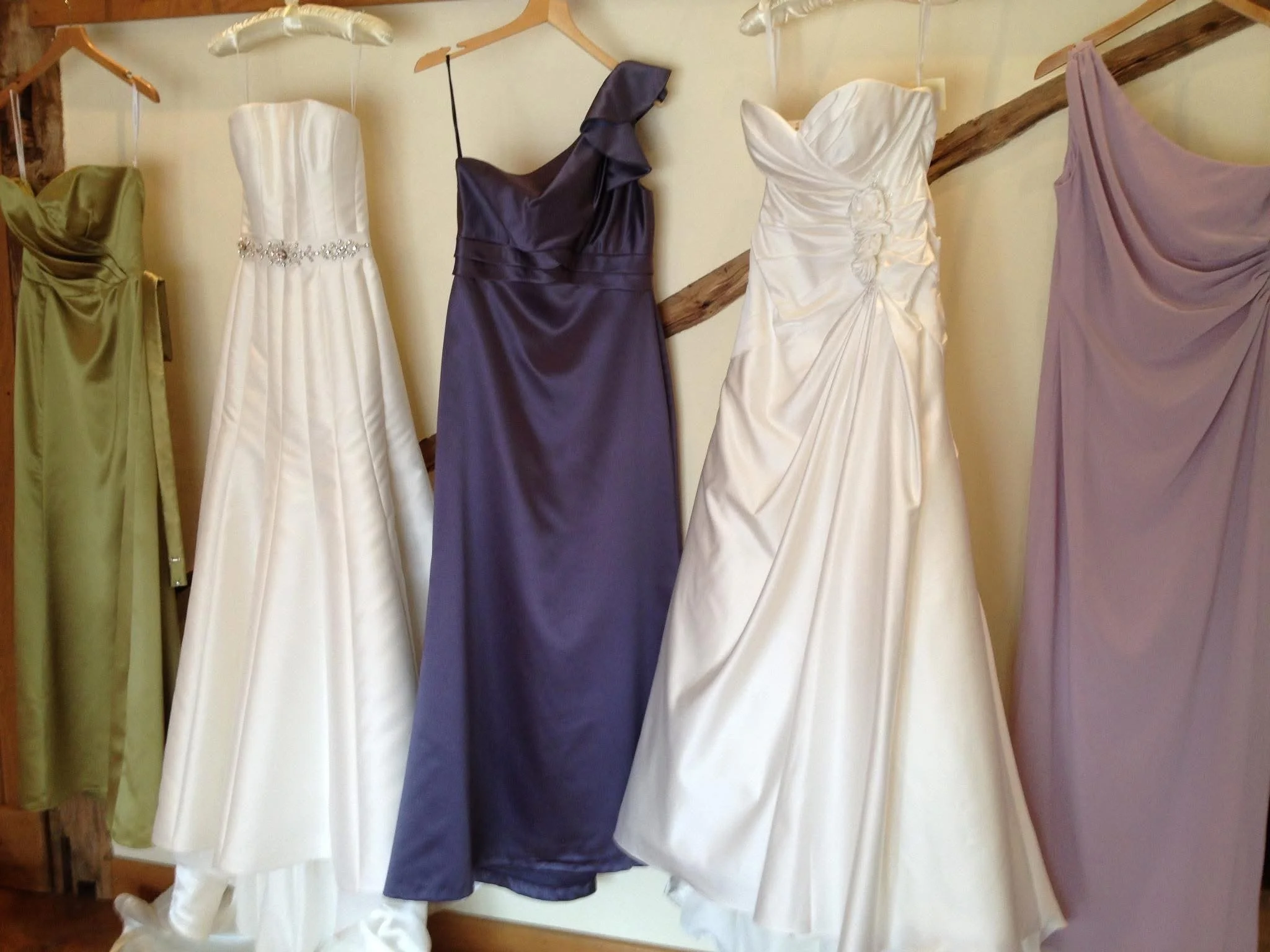 A variety of Brides Maids dresses hanging on a wooden rack. The dresses are in different colors including green, white, purple, cream, and lavender, and some have decorative embellishments.