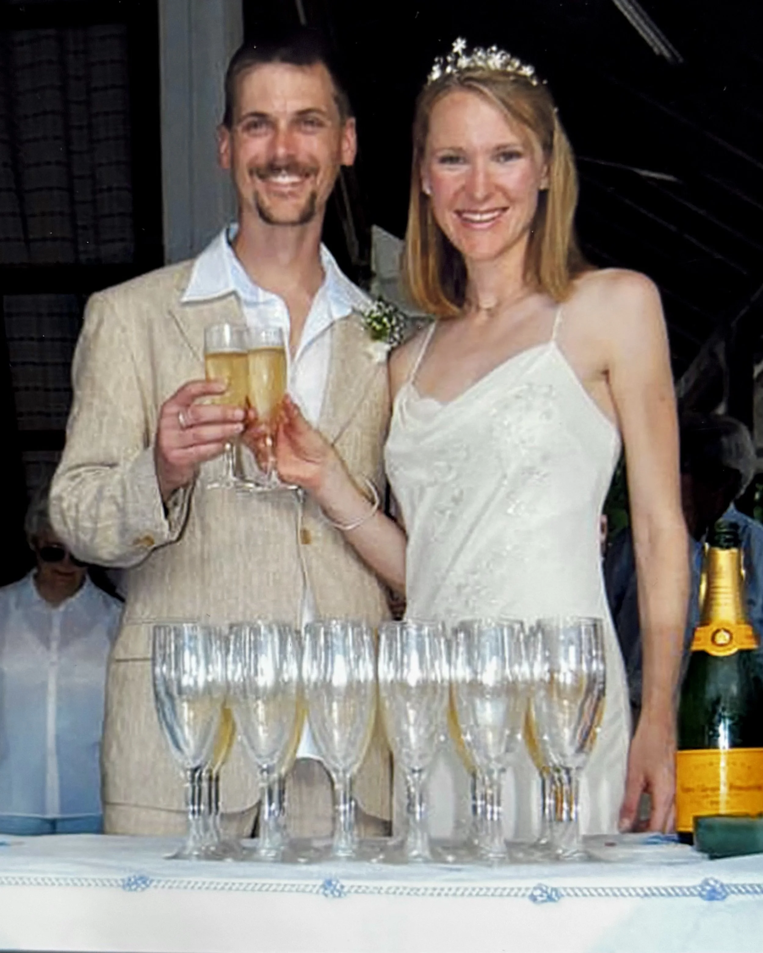 Two people dressed in wedding attire, one man and one woman, holding glasses of champagne, celebrating at a wedding reception table with empty champagne glasses and a yellow beer bottle on the side.