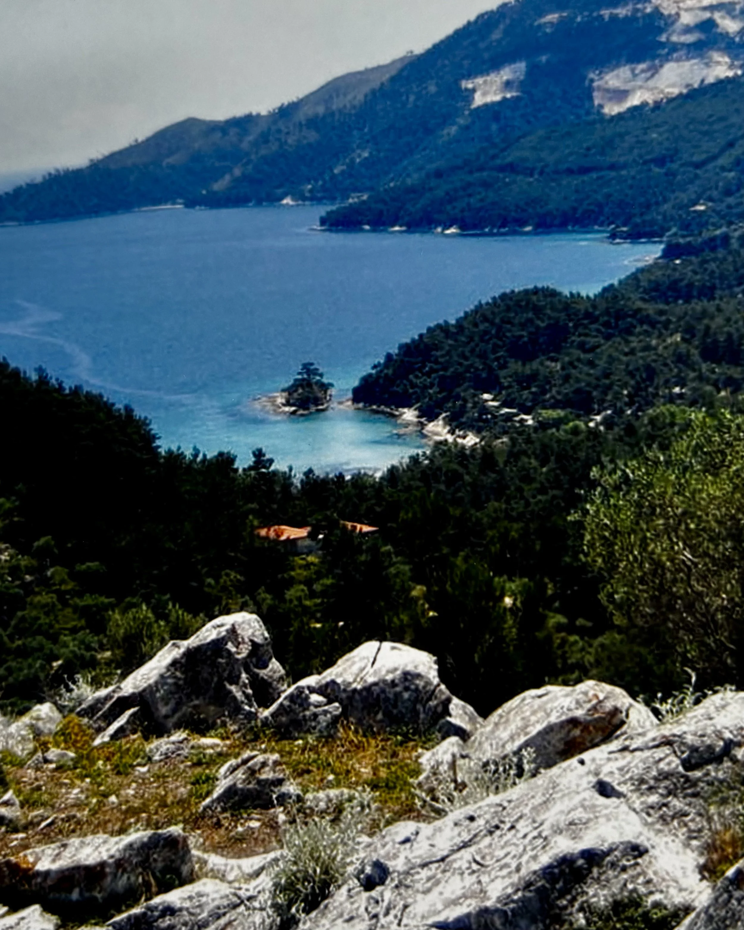 Mountain landscape with a blue sea, forested hills, and rocky foreground under a cloudy sky.