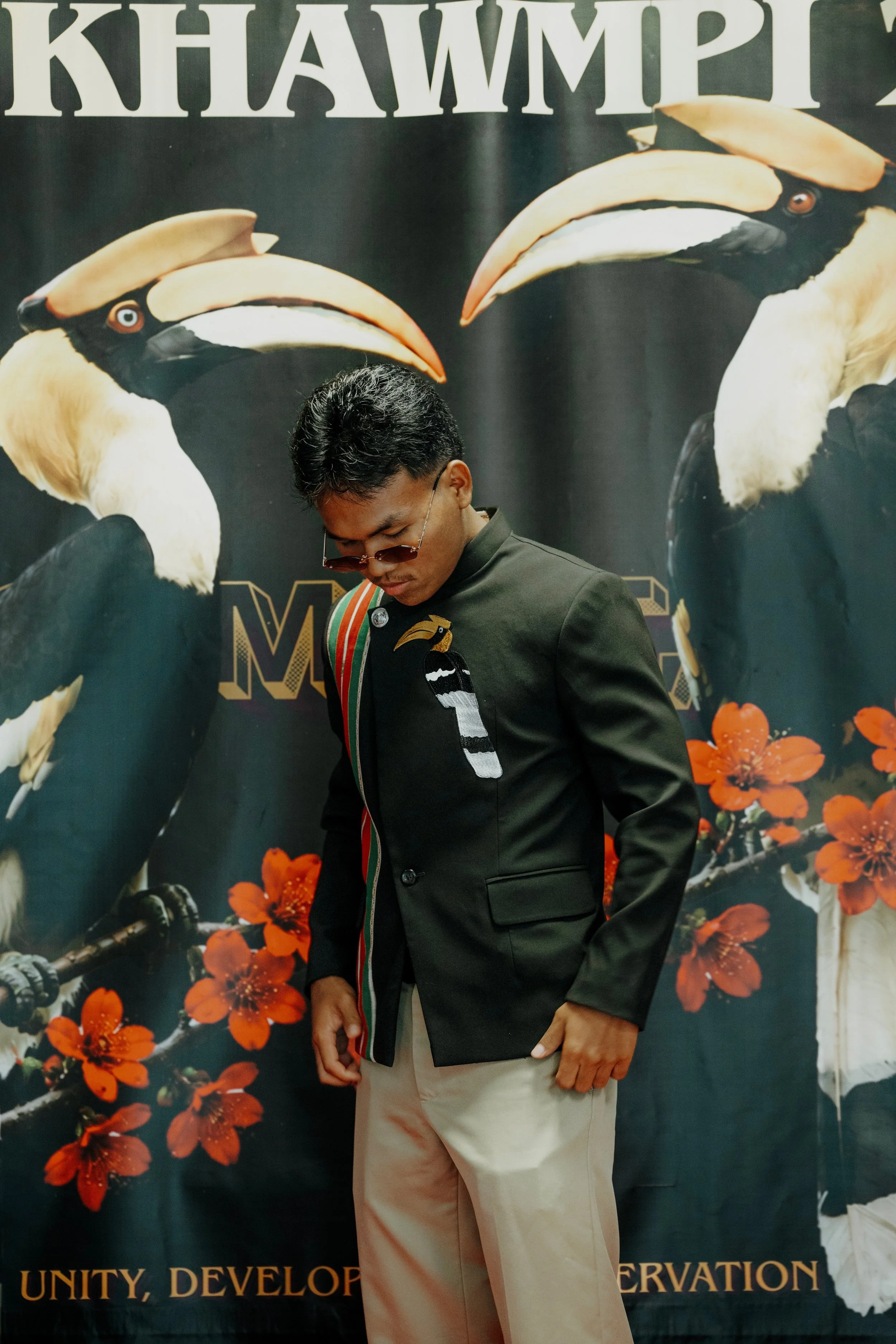 A man wearing glasses, a black jacket with a colorful striped ribbon, and light-colored pants, standing in front of a black backdrop with large images of hornbills and orange flowers. Part of the backdrop includes partial text, likely related to unity, development, and conservation.