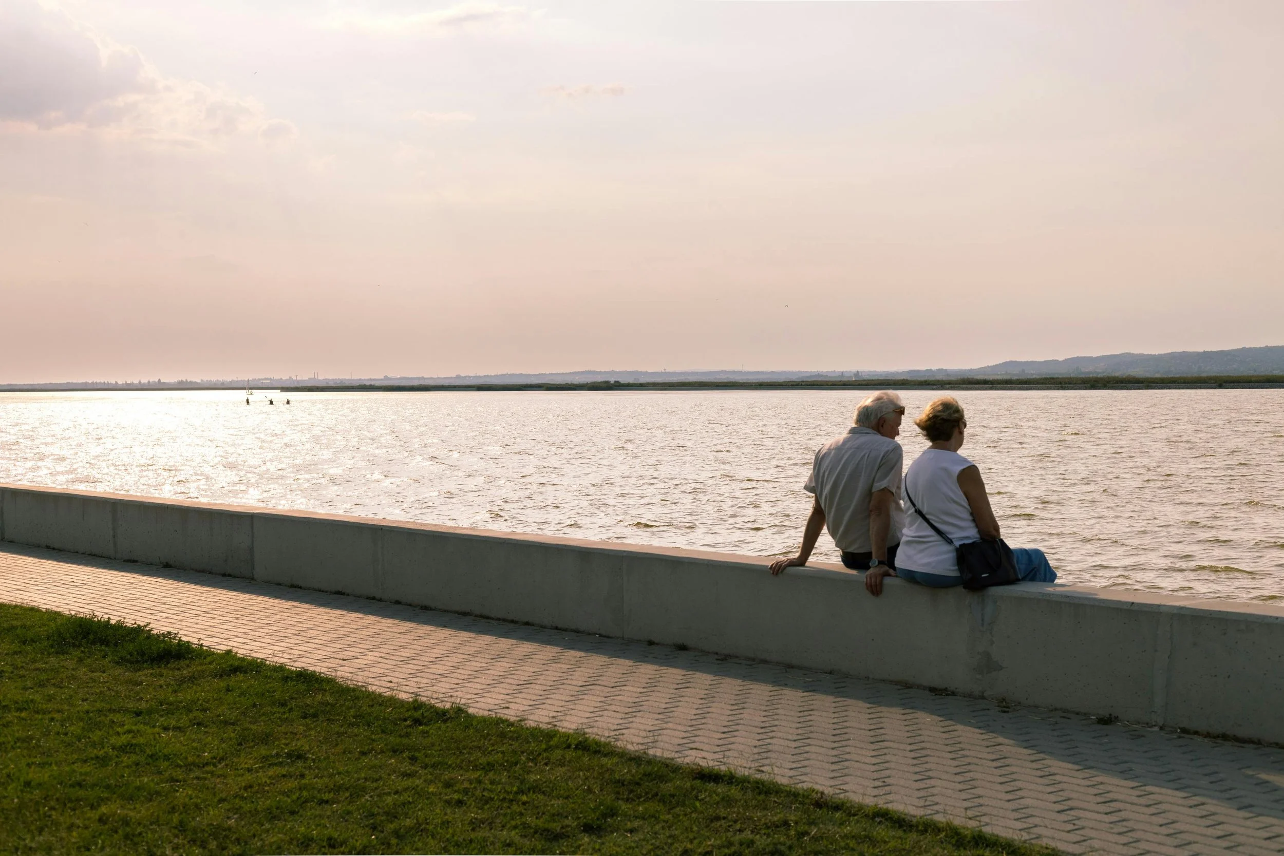 Two elderly people sitting on a concrete barrier by a large body of water, engaging in conversation, with a cloudy sky and distant landscape in the background.