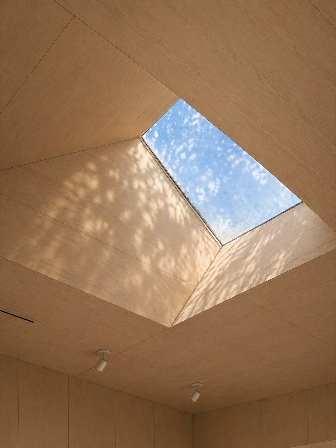 Interior ceiling with a large square skylight showing blue sky and white clouds, wood-paneled ceiling with two ceiling-mounted lights.