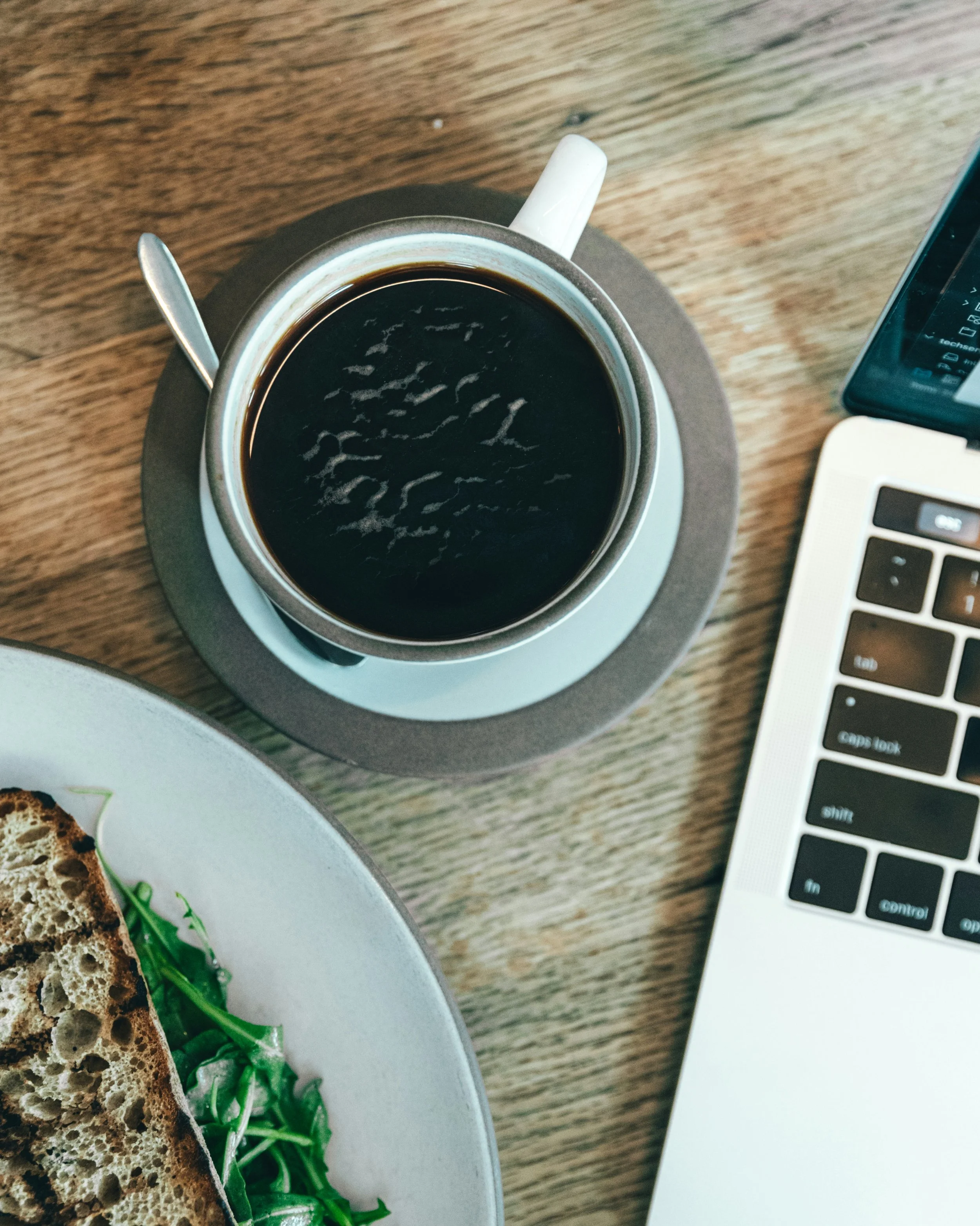 A cup of black coffee sits next to a laptop computer and sandwich. Photo credit: Spencer Davis on Unsplash