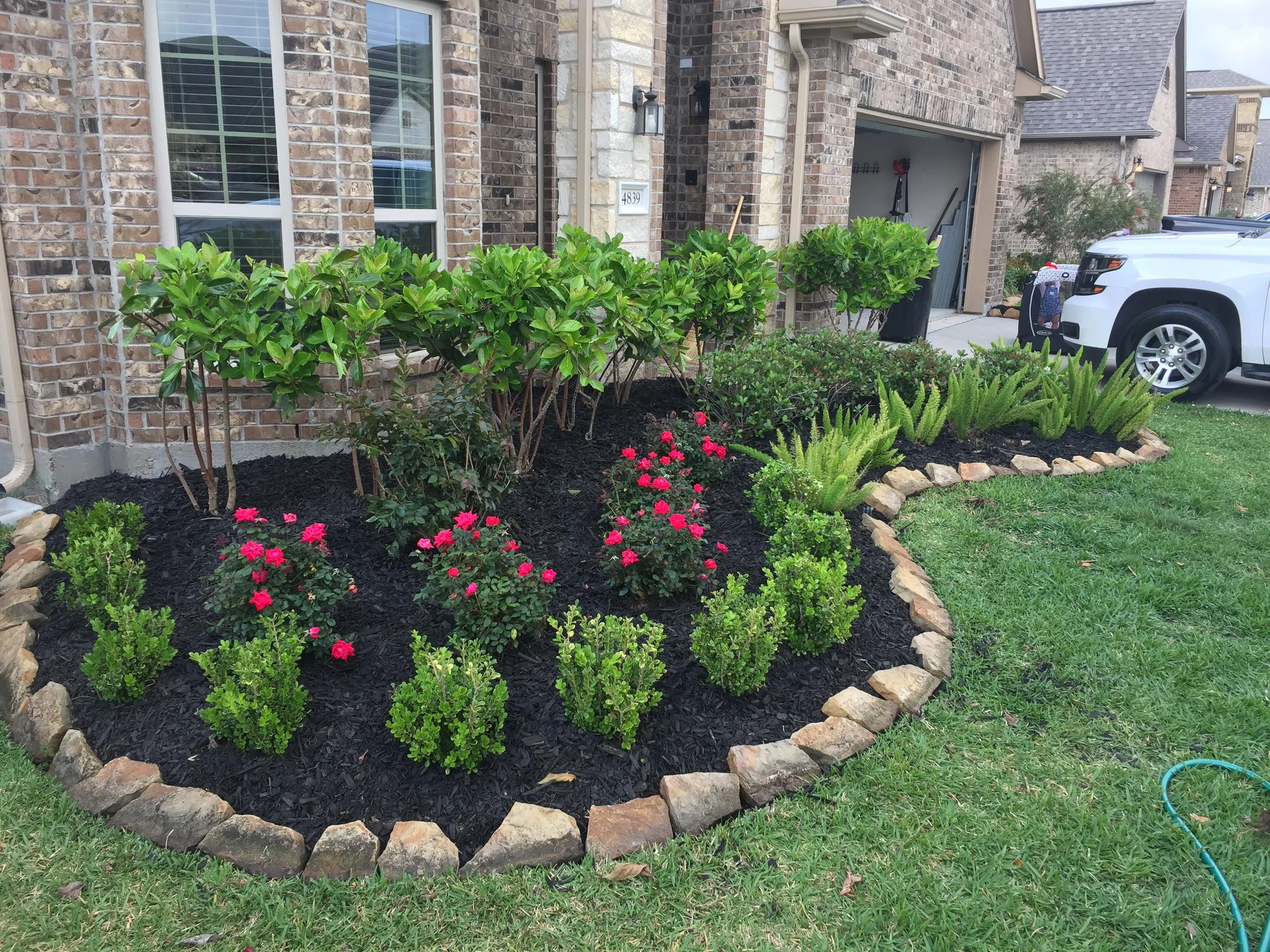 Front yard landscaping featuring a flower bed with pink and red flowers, green shrubs, and small trees, surrounded by a border of rocks. In the background, a brick house with a white garage door and a white vehicle parked outside.