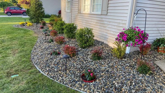 A landscaped garden bed with small bushes and flowering plants, covered with decorative rocks, with black landscape lights and a pink flowering plant in a pot, next to the beige house siding.