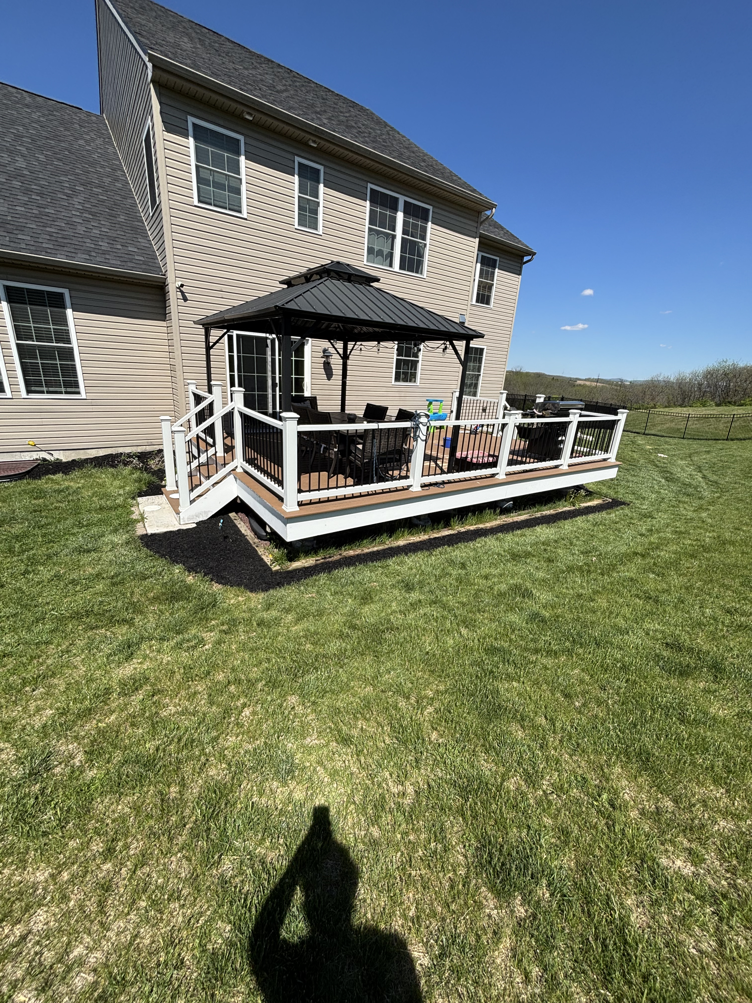 Backyard deck with a black metal gazebo, outdoor furniture, and a grill, next to a beige house with multiple windows against a clear blue sky.