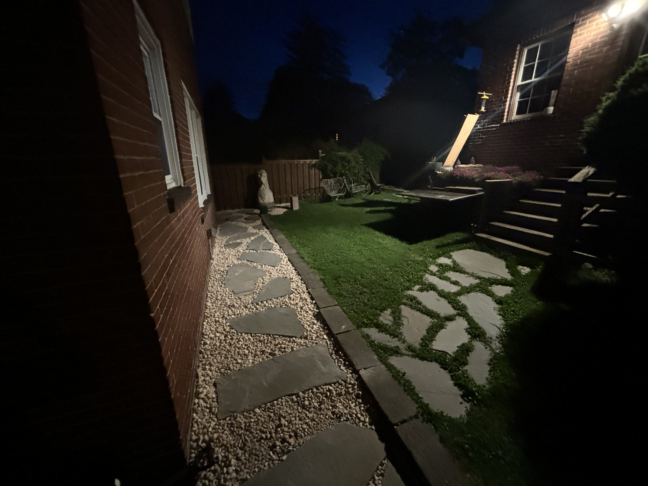 Nighttime backyard with a brick house on the left, a wooden fence, patio furniture, a small garden, and a tree, illuminated by outdoor lights.