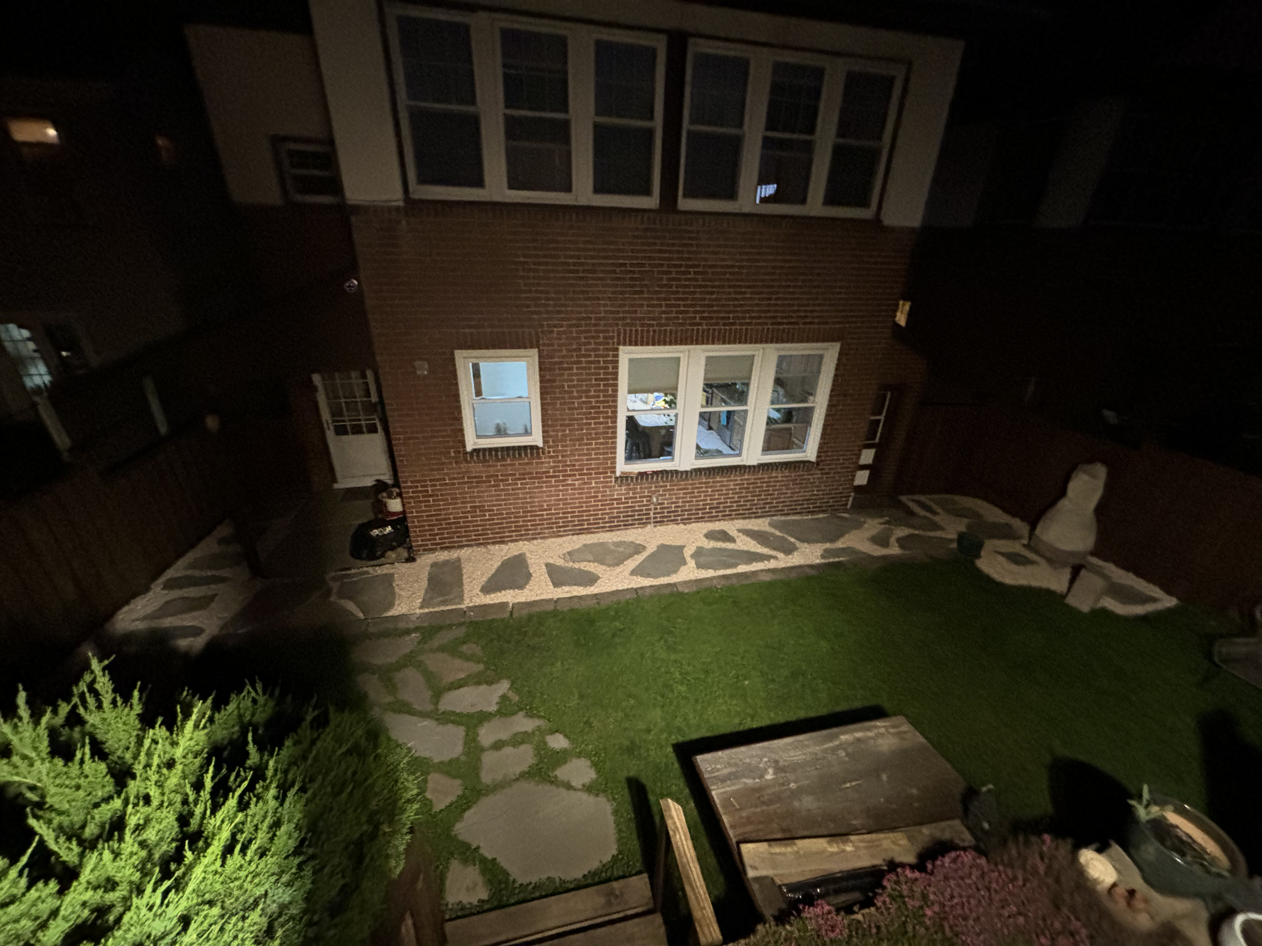 Night view of the backyard showing a brick house with multiple windows, a stone pathway, a green lawn, small bushes, a picnic table, and garden furniture.
