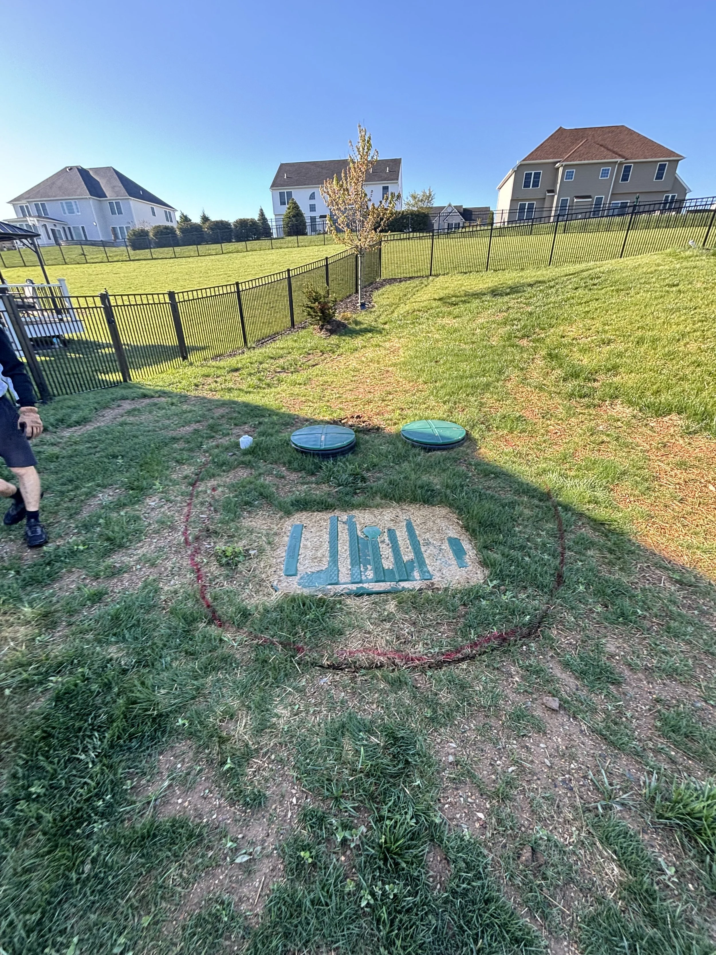 A backyard with green grass, a circular red boundary marking, and two green covered utility access points in the center. There are houses on a hillside in the background under a clear blue sky, and a person partially visible on the left side of the i