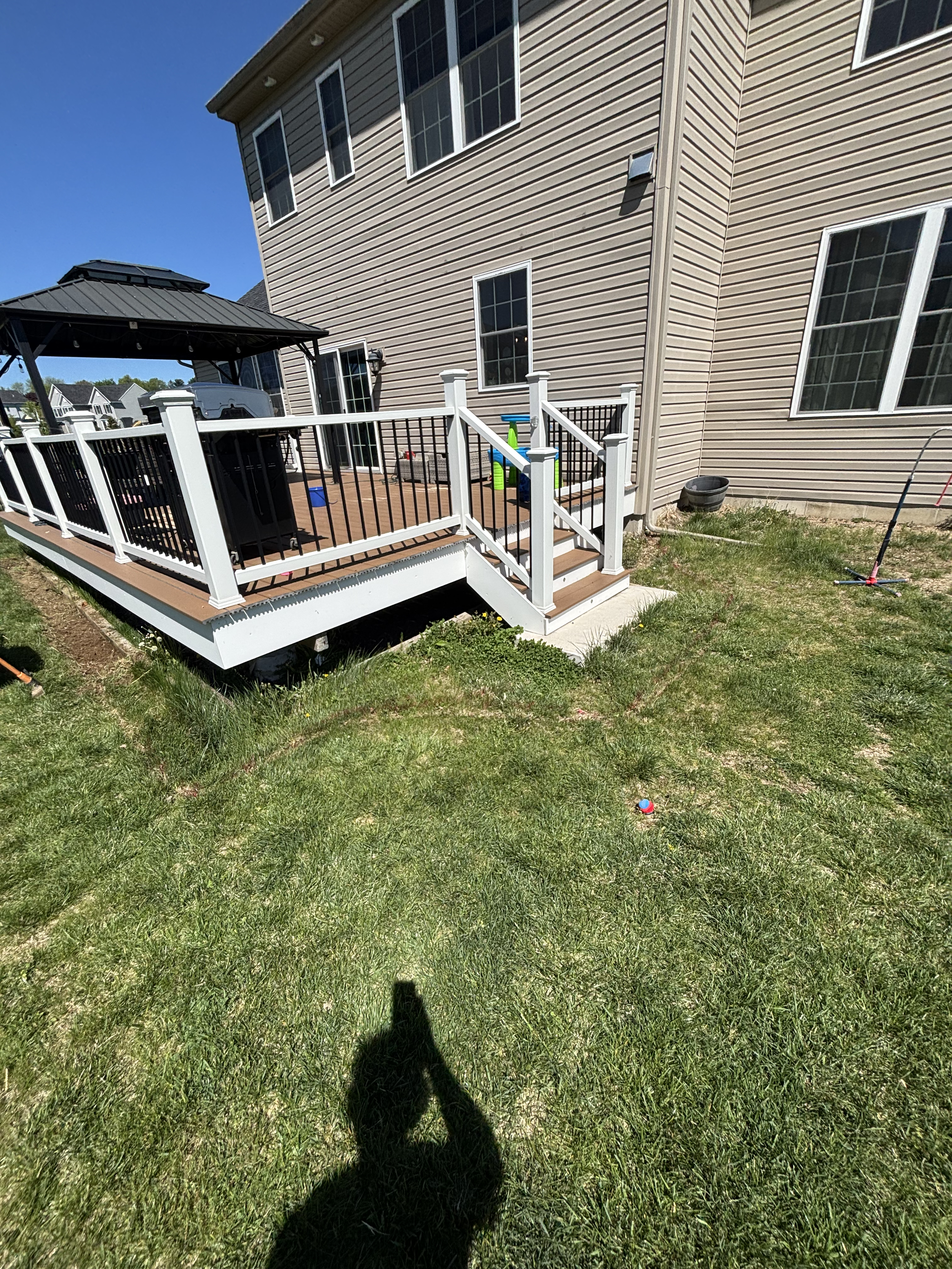 Backyard of a house featuring a wooden deck, white railing with black balusters, a small slide, and a grassy lawn with toys scattered, under a clear blue sky.