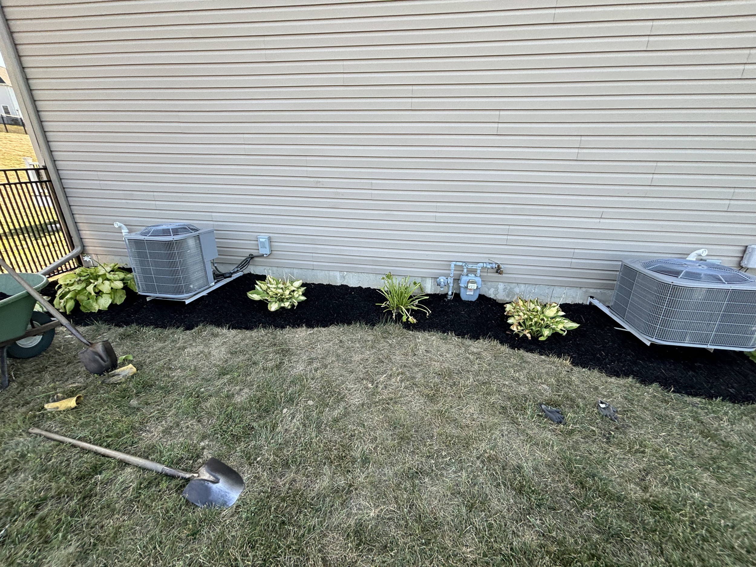 Image of the side of a house with beige siding, two air conditioning units, garden plants, and gardening tools on the grass.