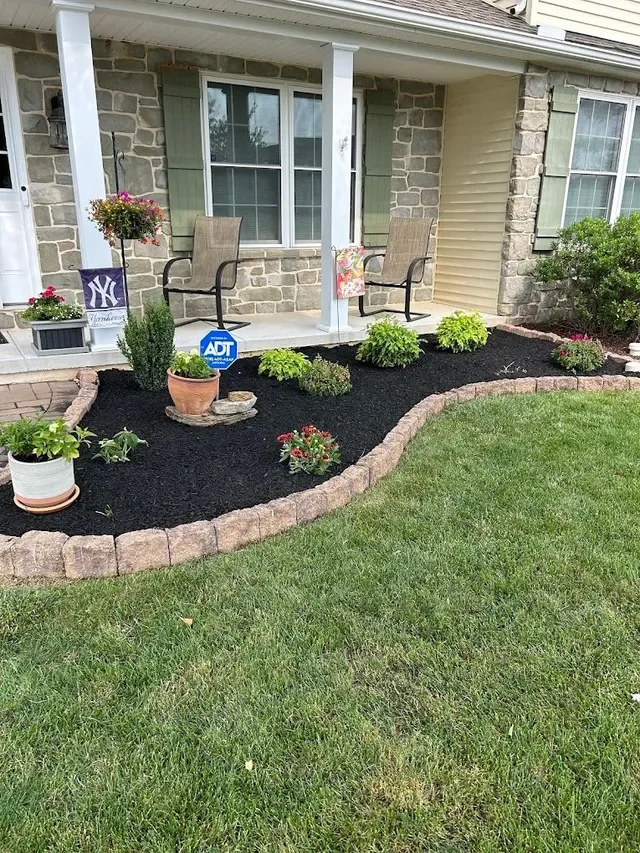 Front yard flower bed with mulch, edged with bricks, containing potted plants, small bushes, and flowers, with two chairs on a porch and a stone and brick house in the background.