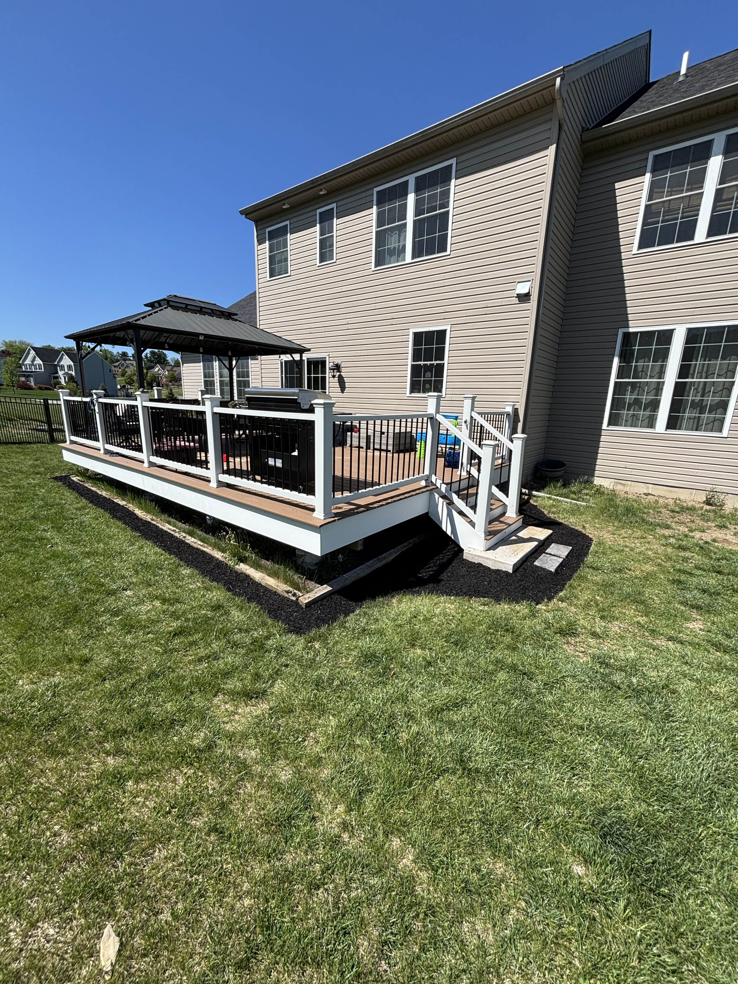 Backyard with a raised wooden deck, black fencing, a gazebo, and a beige house with multiple large windows under a clear blue sky.