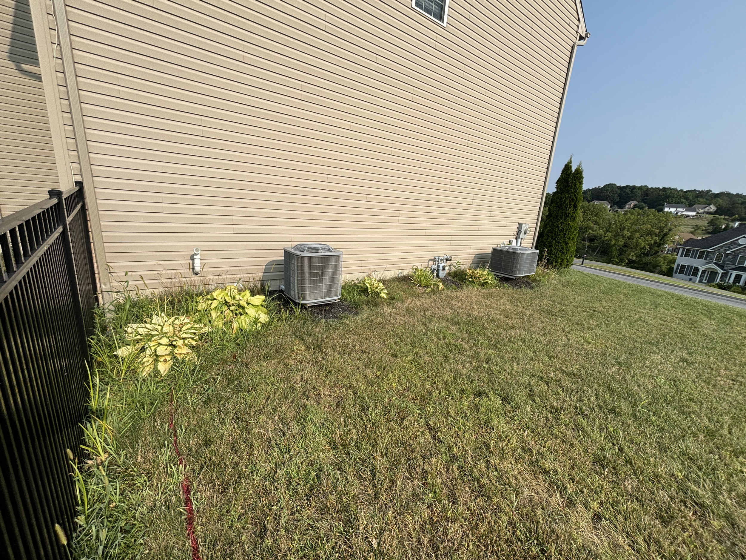 Side view of a beige house with horizontal siding, two air conditioning units on the ground, a white pipe, and small plants in the yard, with other houses and trees in the background under a clear blue sky.
