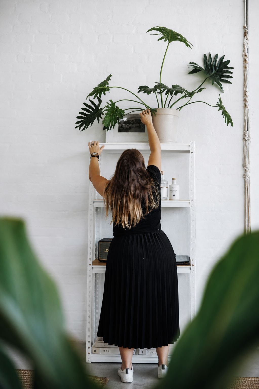Woman with long hair wearing a black dress and sneakers, reaching up to adjust a large potted green plant on a white shelf against a white brick wall.
