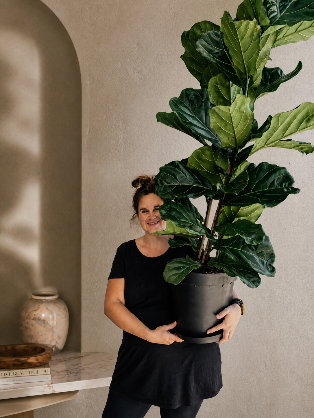 Woman holding a large potted fiddle leaf fig plant indoors, standing near a beige wall with decorative items on a marble table.