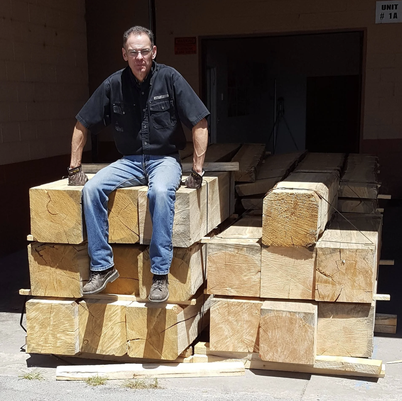 A man sitting on stacked cut wooden logs at a lumber yard, wearing a black shirt and jeans.