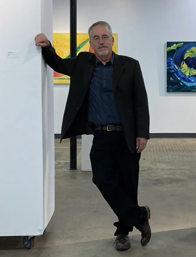 A man with gray hair and glasses posing in an art gallery, standing with one arm resting on a white wall partition, wearing a black blazer, dark blue shirt, black pants, and brown shoes, with colorful abstract paintings on the wall behind him.