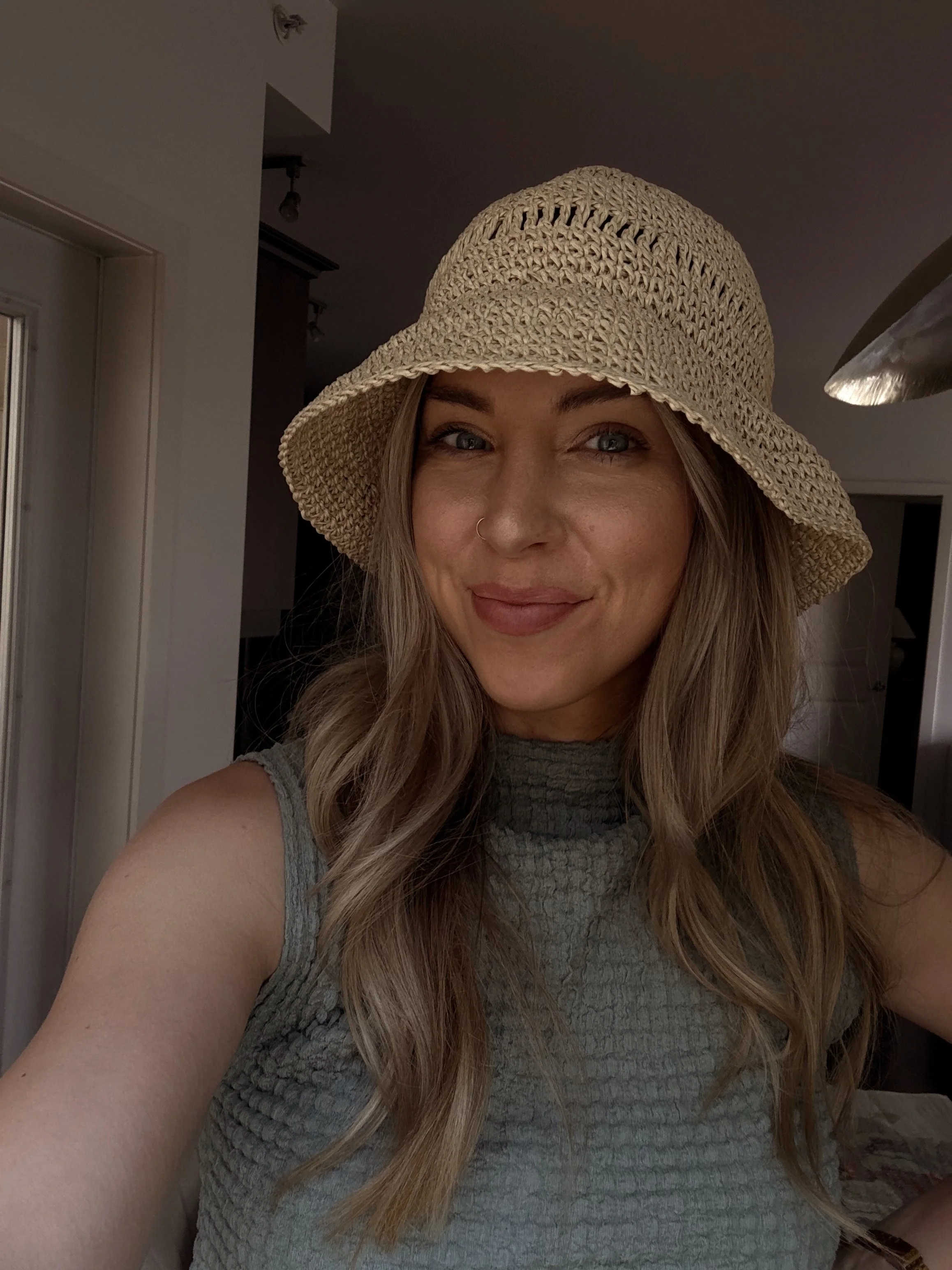 A woman with long wavy hair, wearing a beige knitted hat and a sleeveless, textured gray top, smiling in an indoor setting.