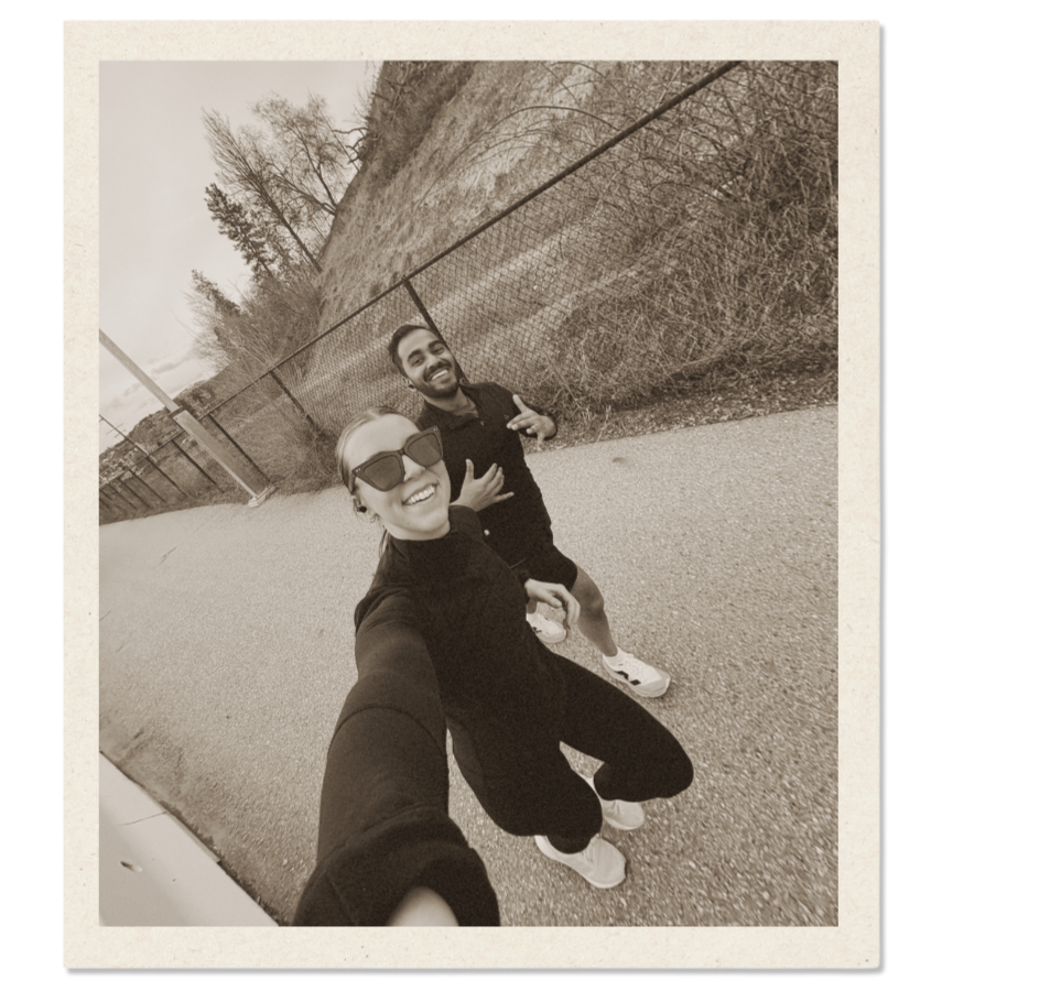 Two smiling people taking a selfie outdoors, with a chain-link fence and trees in the background, in black and white.