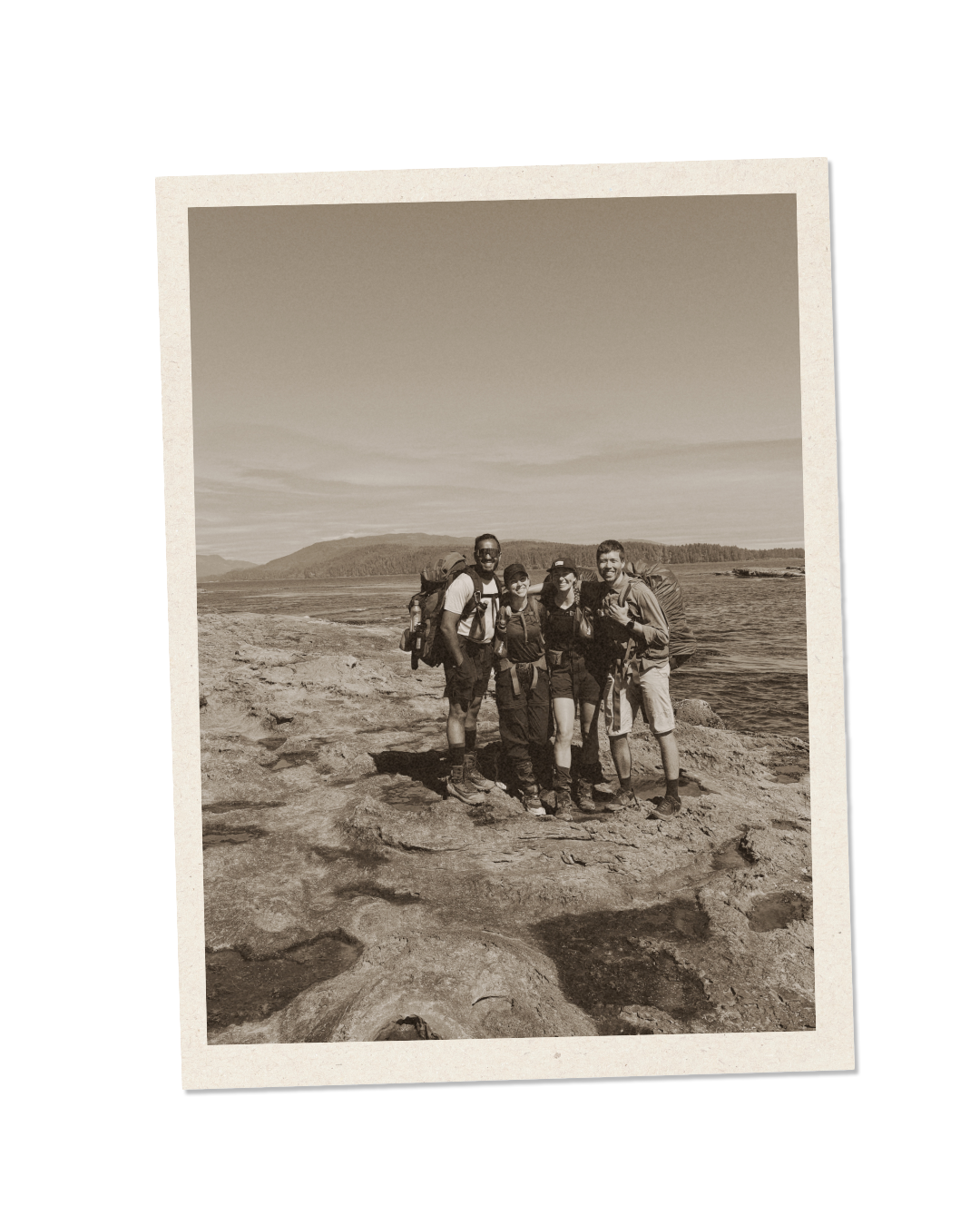 Four people standing outdoors on rocky terrain near water with mountains in the background, wearing hiking gear and backpacks, in black and white.