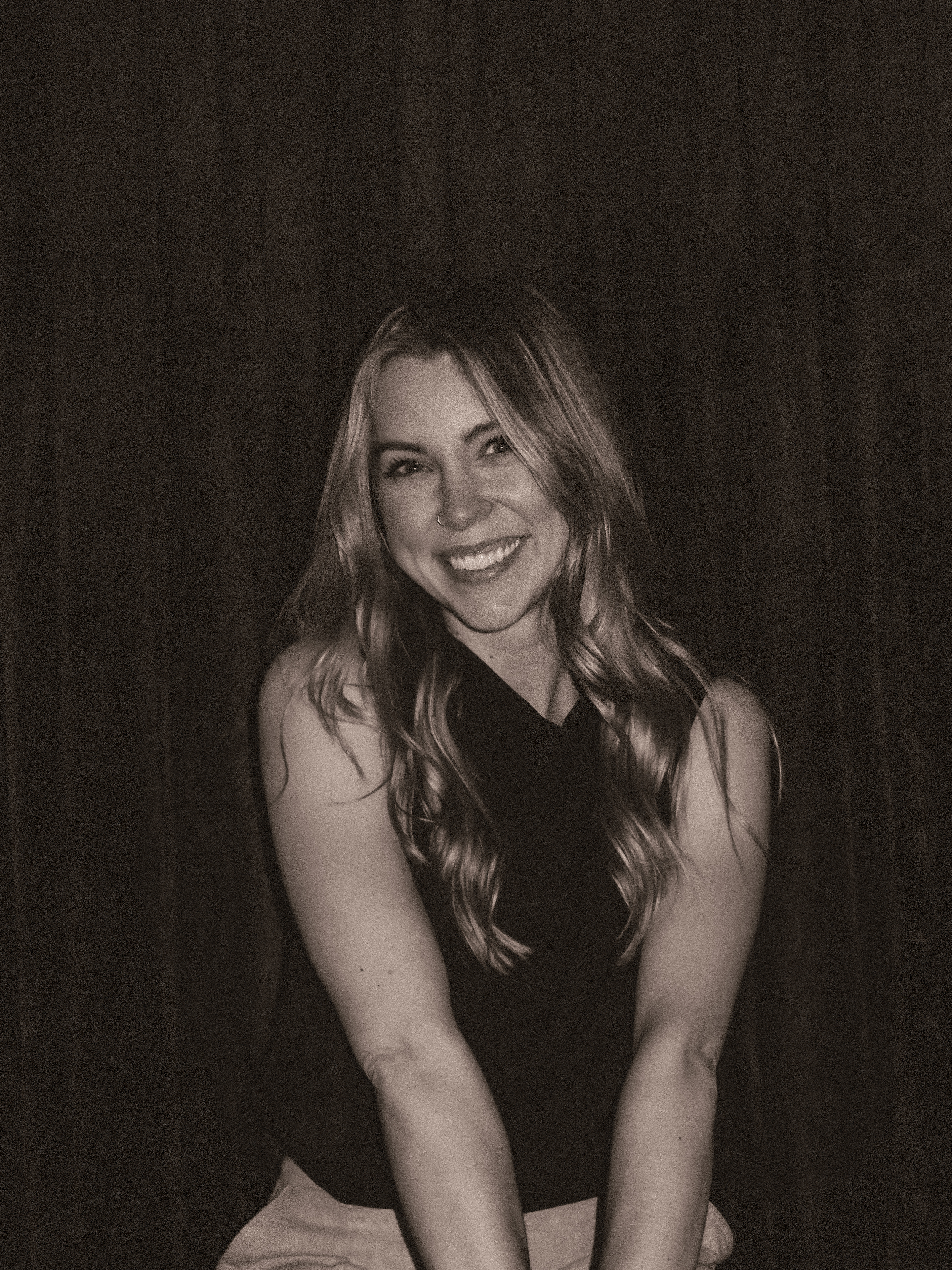 A smiling young woman with long, wavy hair, wearing a nose ring, black top, and light-colored pants, sitting against a dark background.