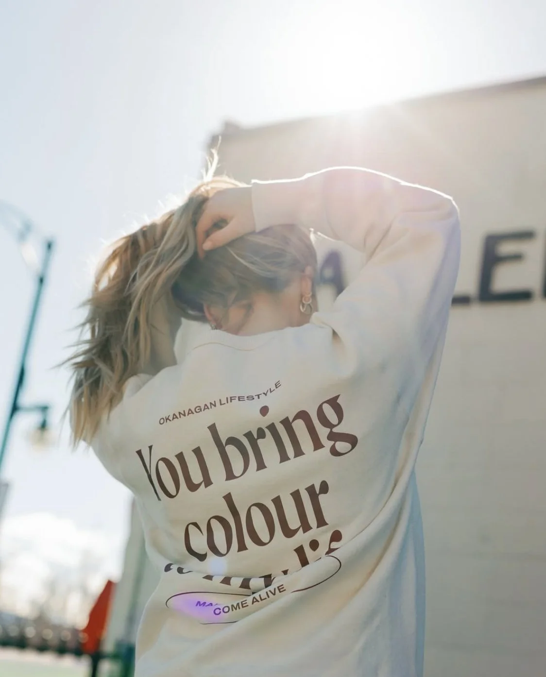 A woman with blonde hair wearing a white sweatshirt with text, standing outdoors, with bright sunlight shining from behind.