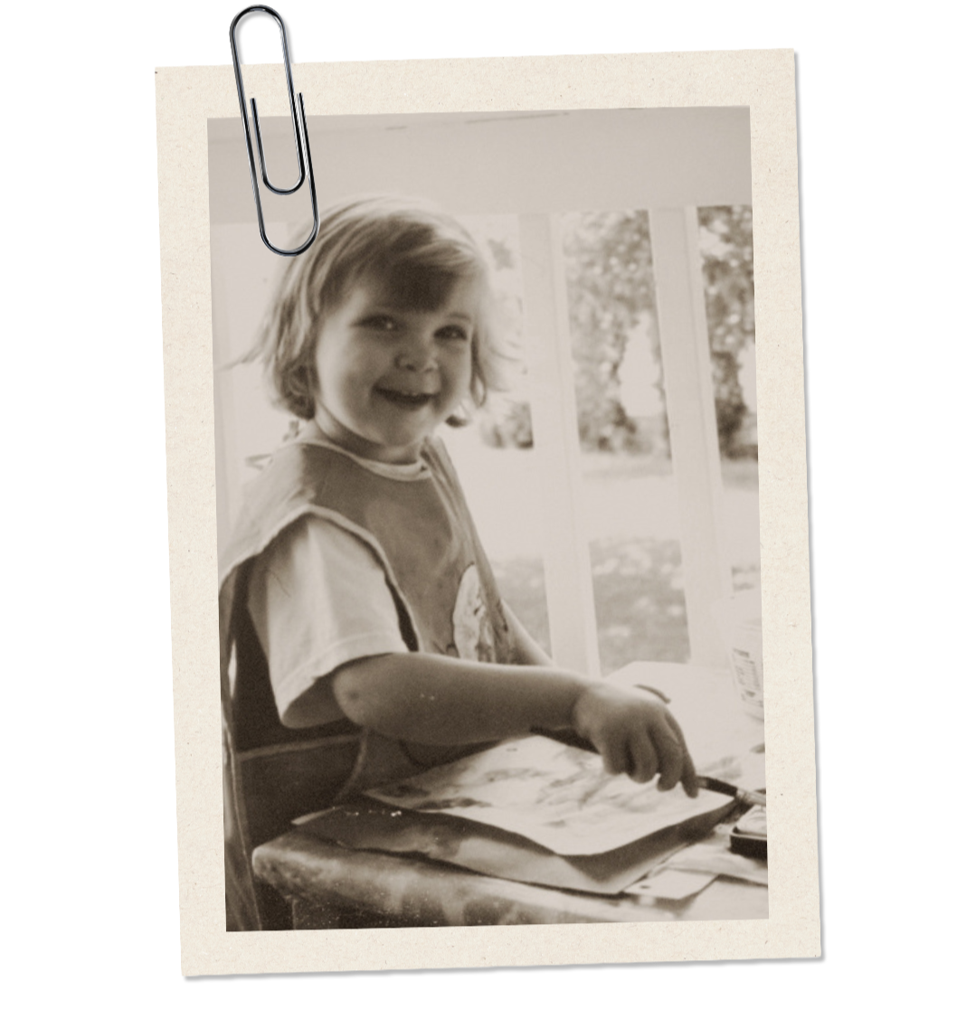 A young girl with shoulder-length hair is smiling and painting at a table, with a window and outdoor scenery in the background.