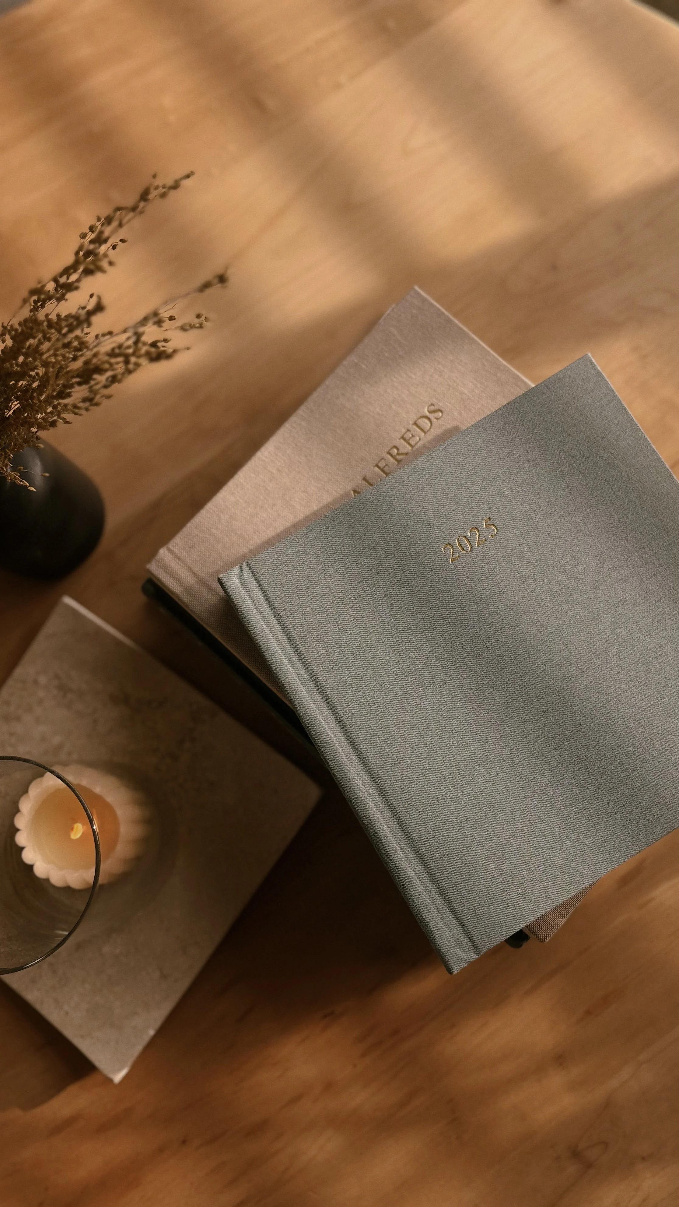 A photograph of two journals stacked on a wooden table, with the top journal marked '2025' in gold lettering and the one underneath labeled 'PLANNERS'. To the left, there is a black vase with dried flowers, a lit candle in a scalloped glass holder, and a beige-colored stone block.