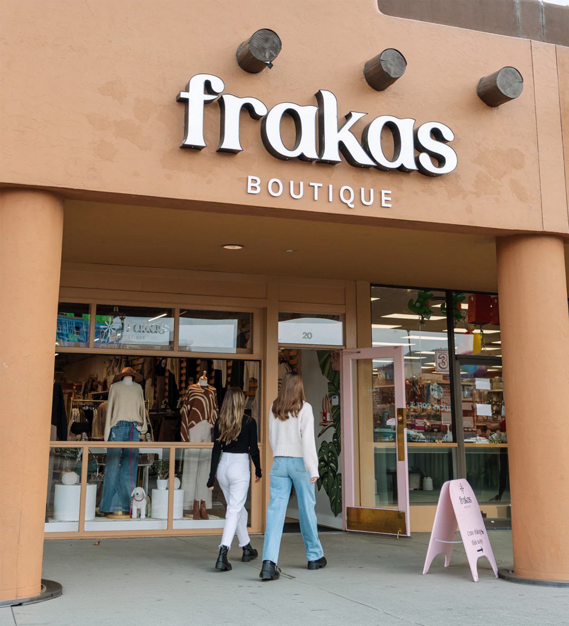 Three women with long hair walk into a boutique clothing store with mannequins and clothing displays visible through the large glass windows and door. The store has a soft peach-colored exterior with rounded columns and a pink signboard outside.