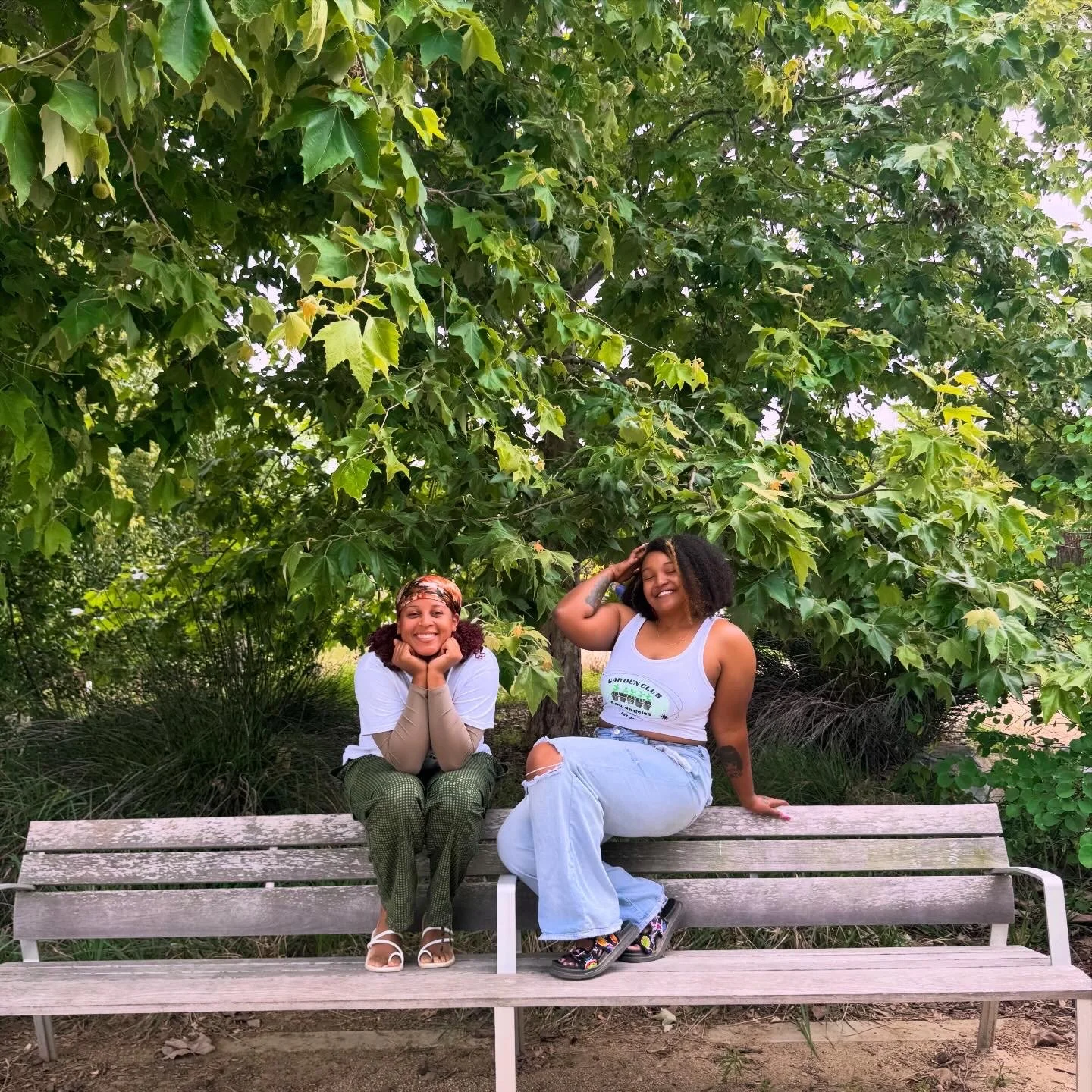 Two women sitting on a park bench under a large leafy tree. One woman is smiling with her chin resting on her hands, and the other woman is smiling and touching her hair. They are outdoors surrounded by greenery.