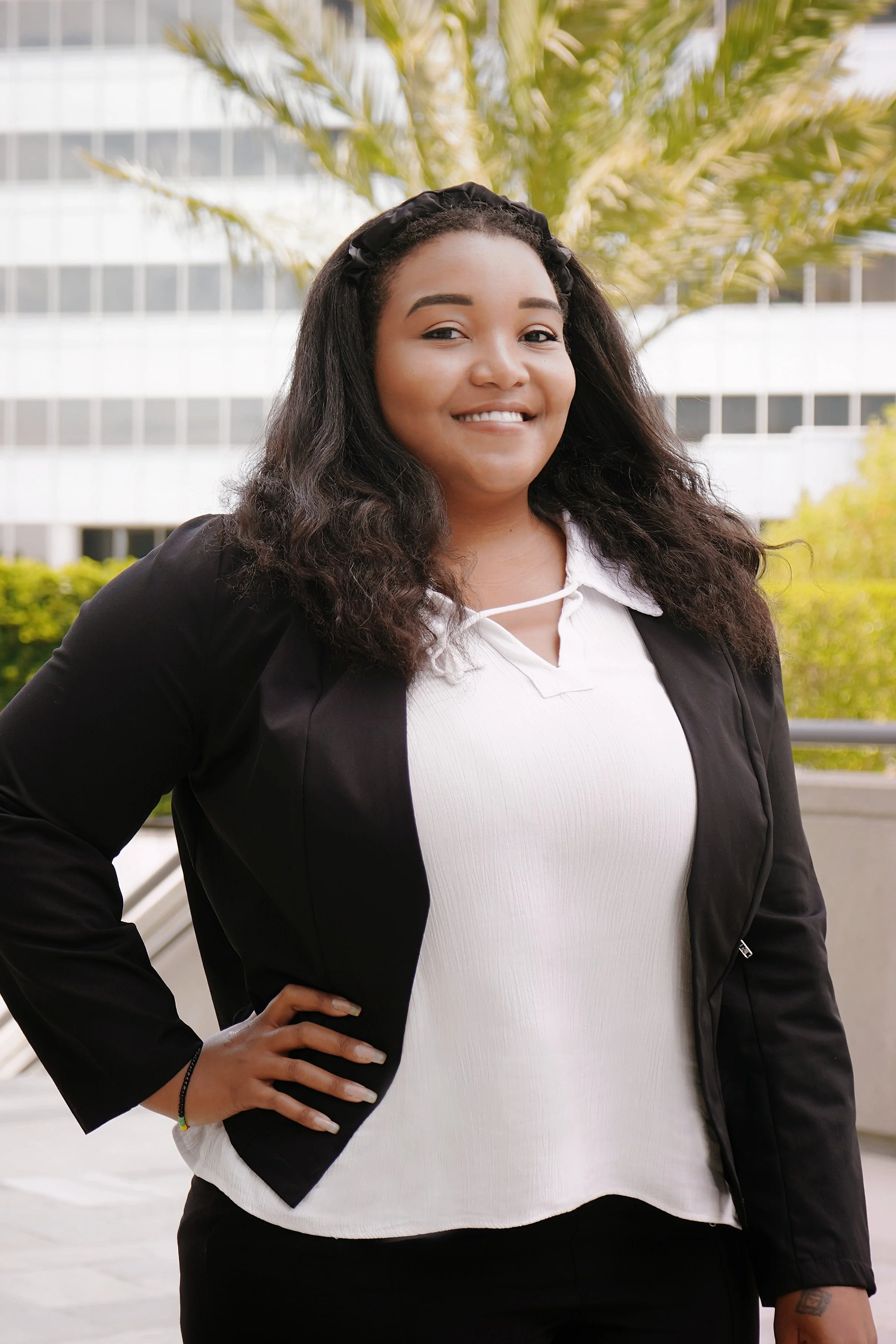 Young woman with curly hair smiling, wearing a black blazer and white shirt, standing outdoors with a building and palm tree in the background.