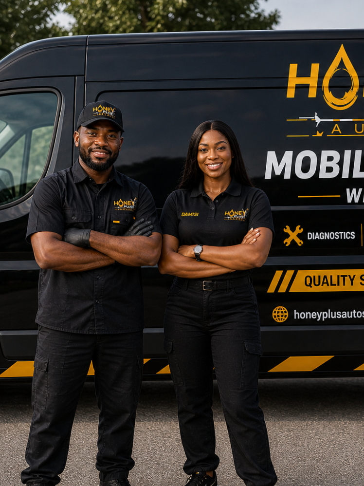 Two auto technicians standing in front of a black service van with yellow and white branding. The man is wearing a black cap and shirt with the company logo, and the woman is wearing a black shirt with her name 'Damisi' on it, both are smiling with arms crossed.