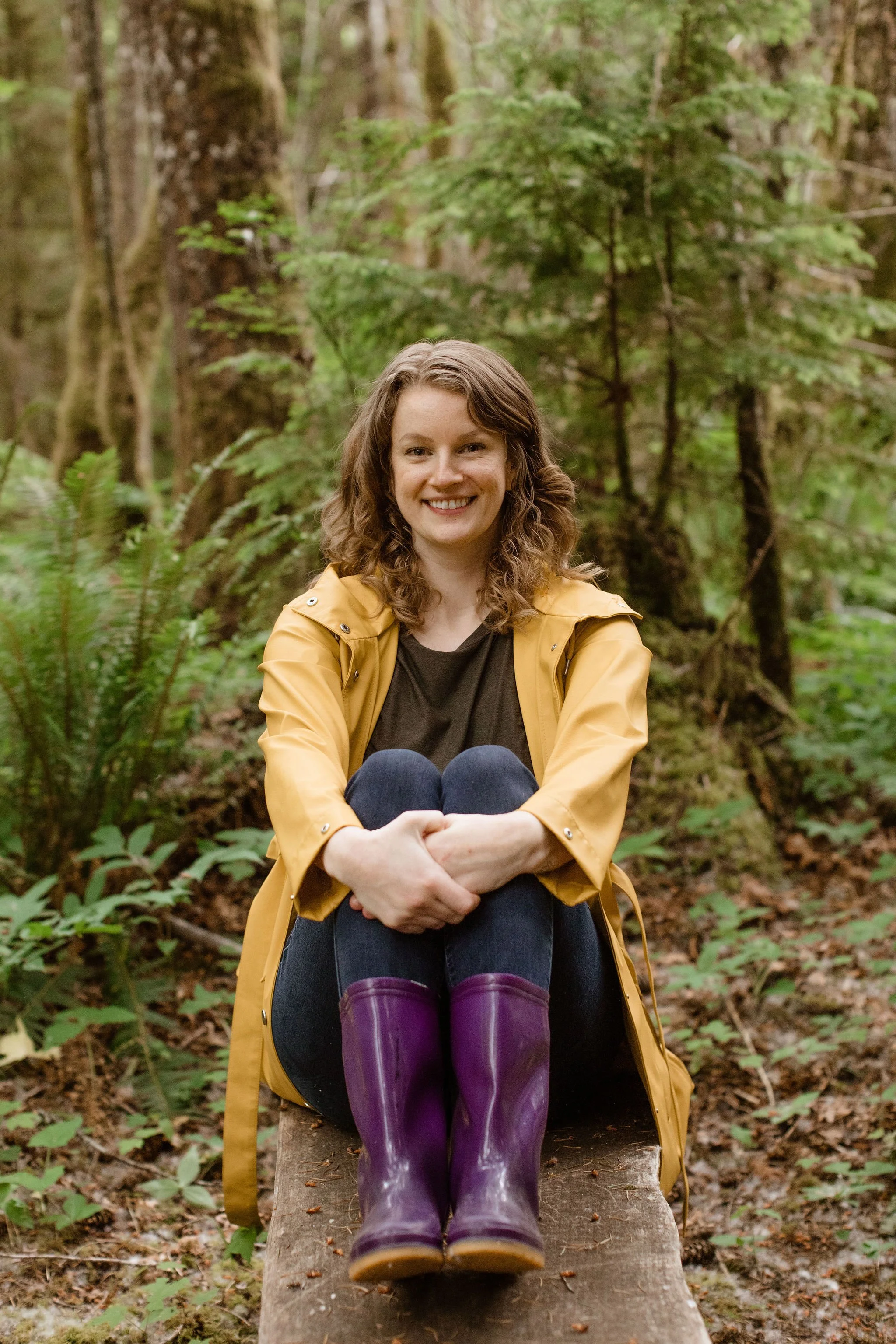 A woman with curly hair, wearing a yellow raincoat, black shirt, blue jeans, and purple rain boots, sitting on a wooden log with her knees to her chest and smiling, in a temperate rainforest surrounded by trees, ferns, and foliage.