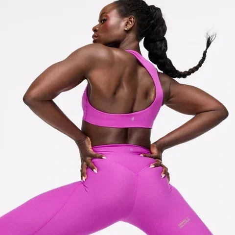 Peloton X Tunde Apparel Collection-A woman in a bright pink sports outfit, standing with her back to the camera, hands on hips, and looking over her shoulder.