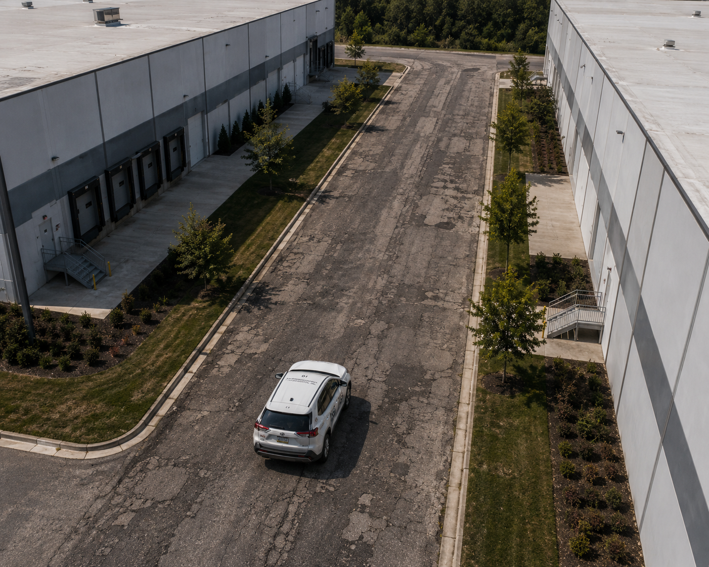Empty parking lot with a white police vehicle parked near a large industrial building. Small trees and shrubs line the sidewalk along the building. The pavement is cracked and worn.