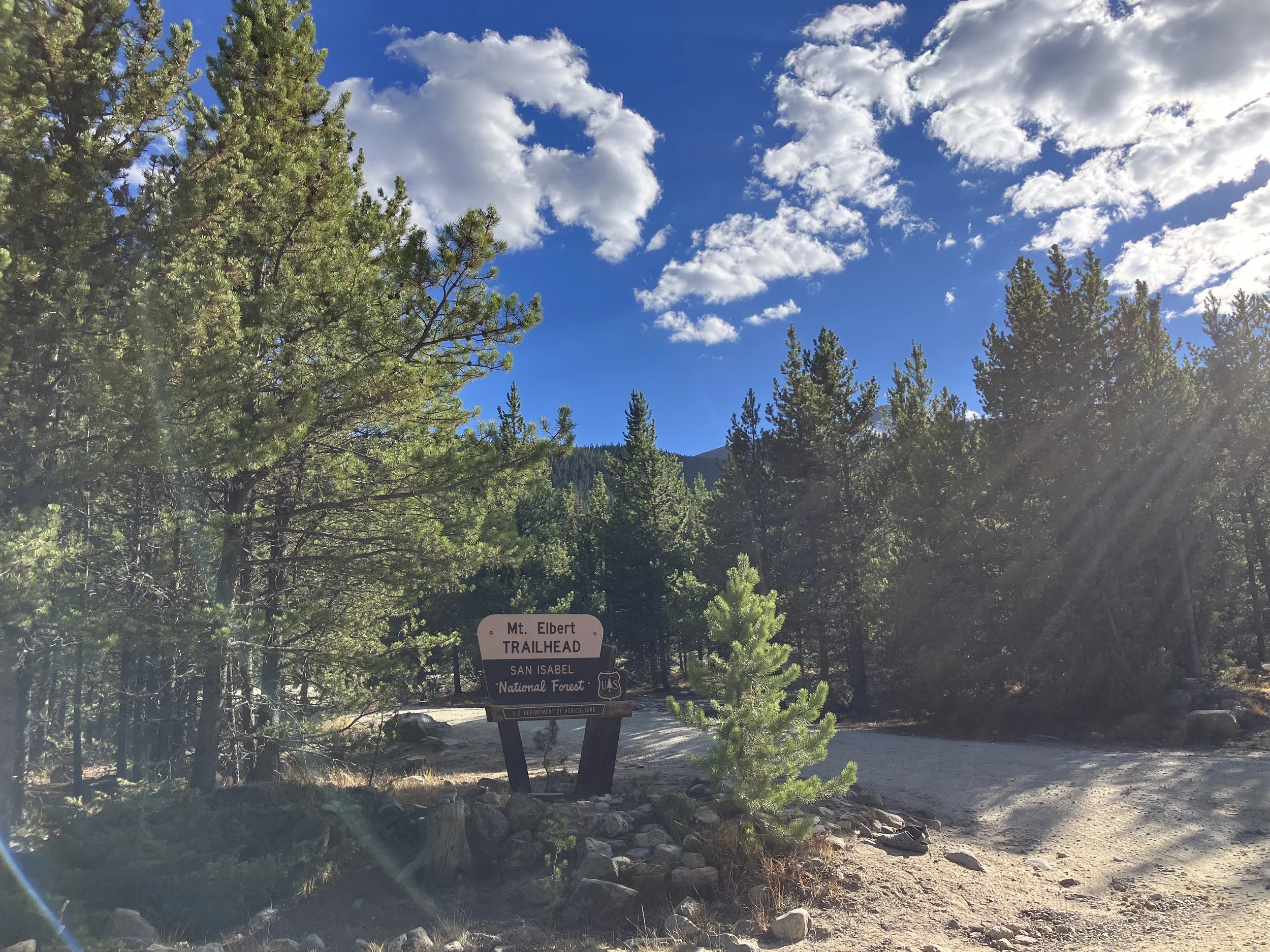 A trailhead sign at Mount Elbert in San Isabel National Forest, surrounded by tall green trees under a partly cloudy blue sky.