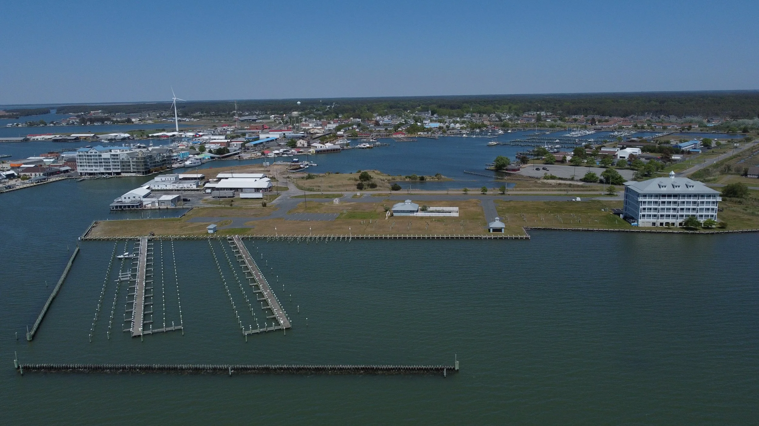 Aerial view of a marina with numerous docks and boats, surrounded by a town with various buildings, water bodies, and greenery under a clear blue sky.