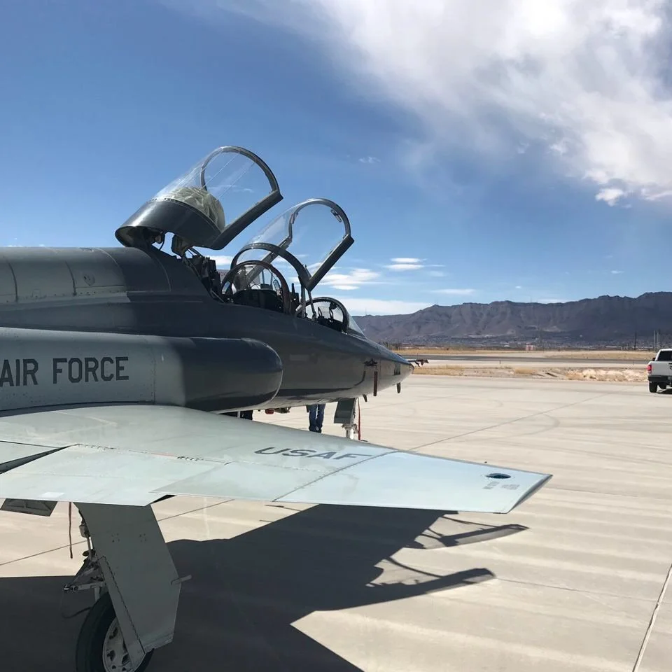 A military jet aircraft with open cockpit canopies parked on an airstrip, with mountains in the background and a clear blue sky.