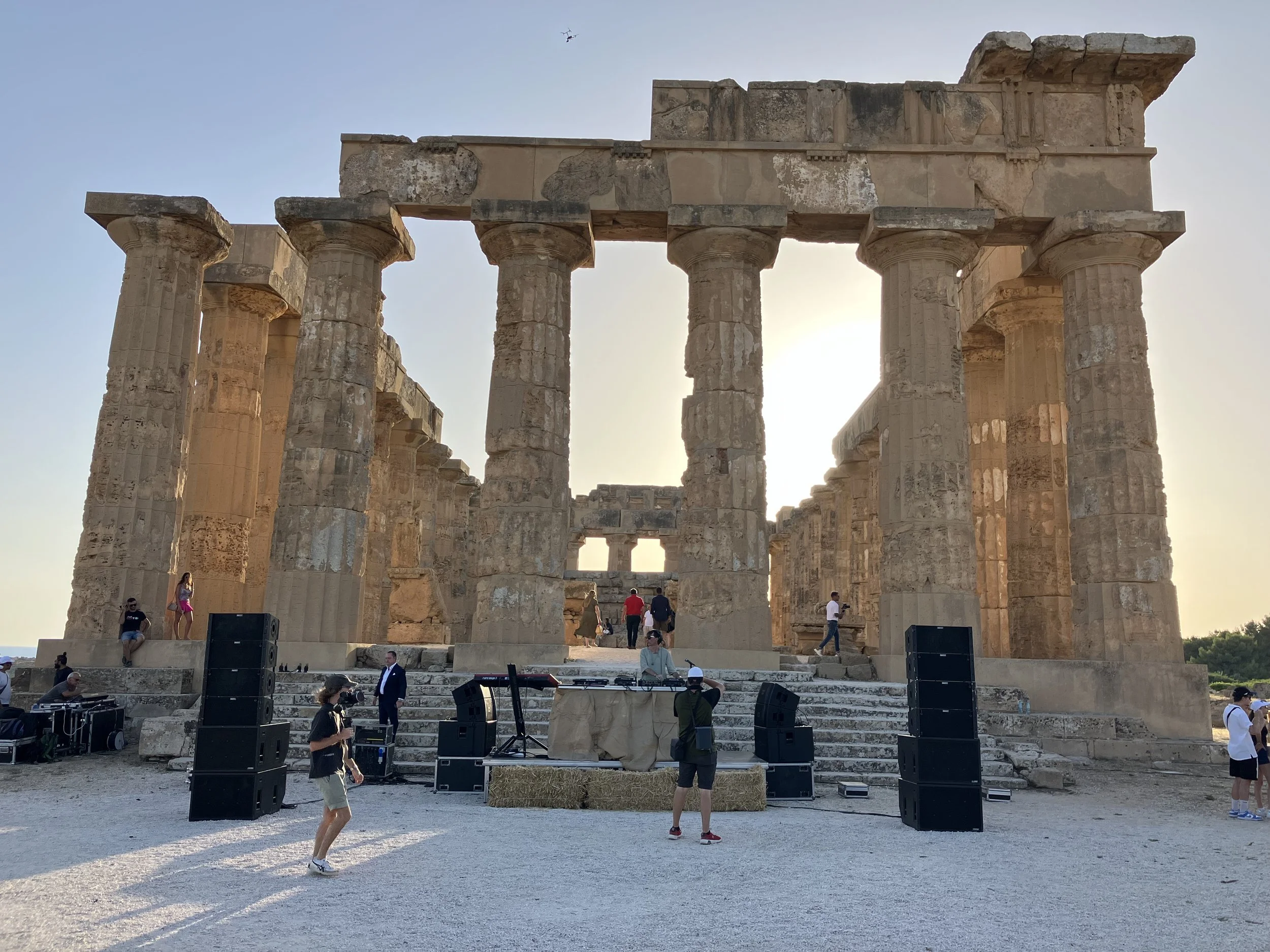 Ancient Greek temple ruins with tall columns, set against a clear sky with the sun low on the horizon. There are people walking and gathered around, some taking photos and others socializing, and sound equipment is set up at the base of the structure