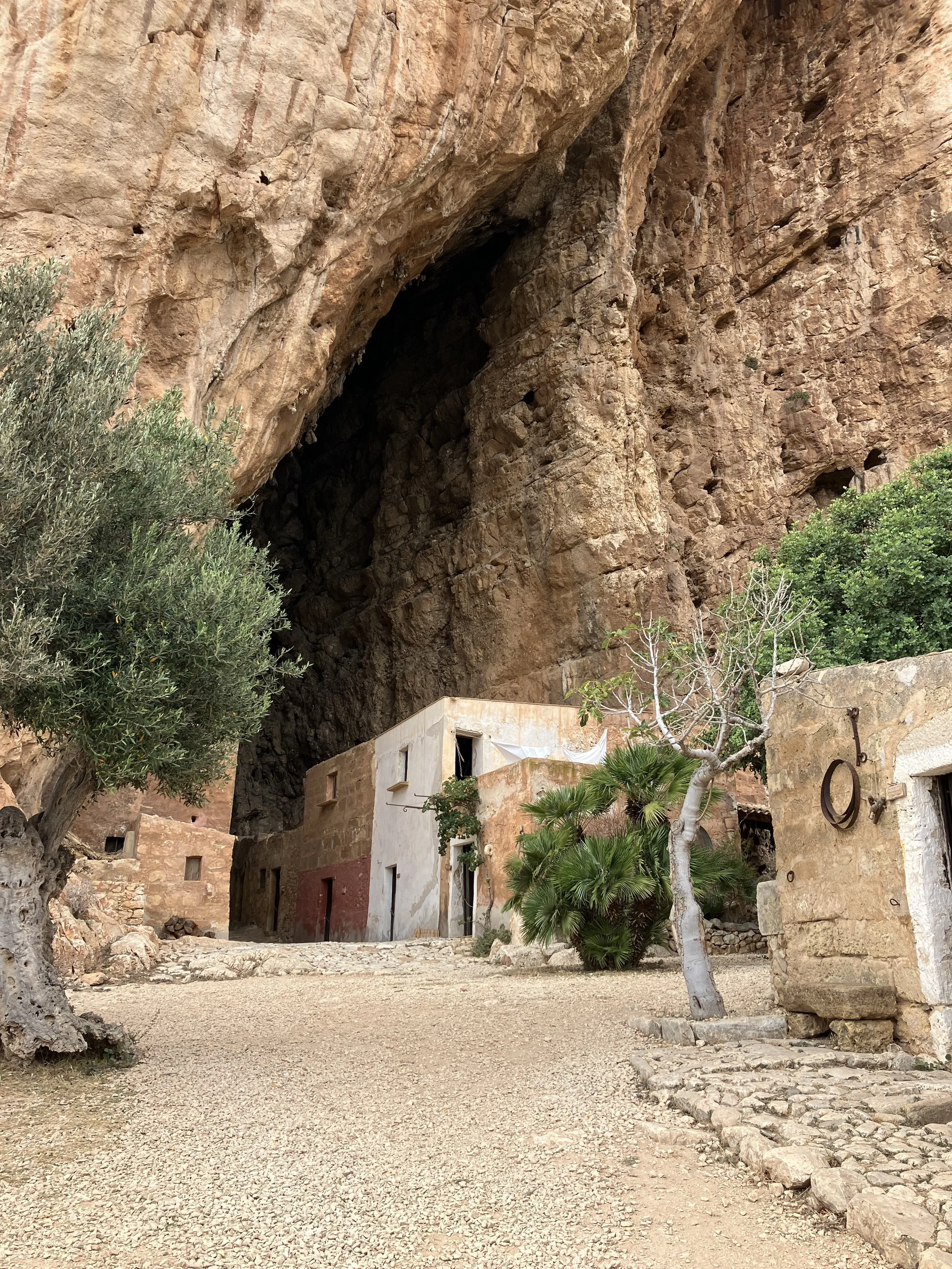 A small indigenous-style village built into a rocky hillside with a large cave opening above, surrounded by desert vegetation and simple structures.