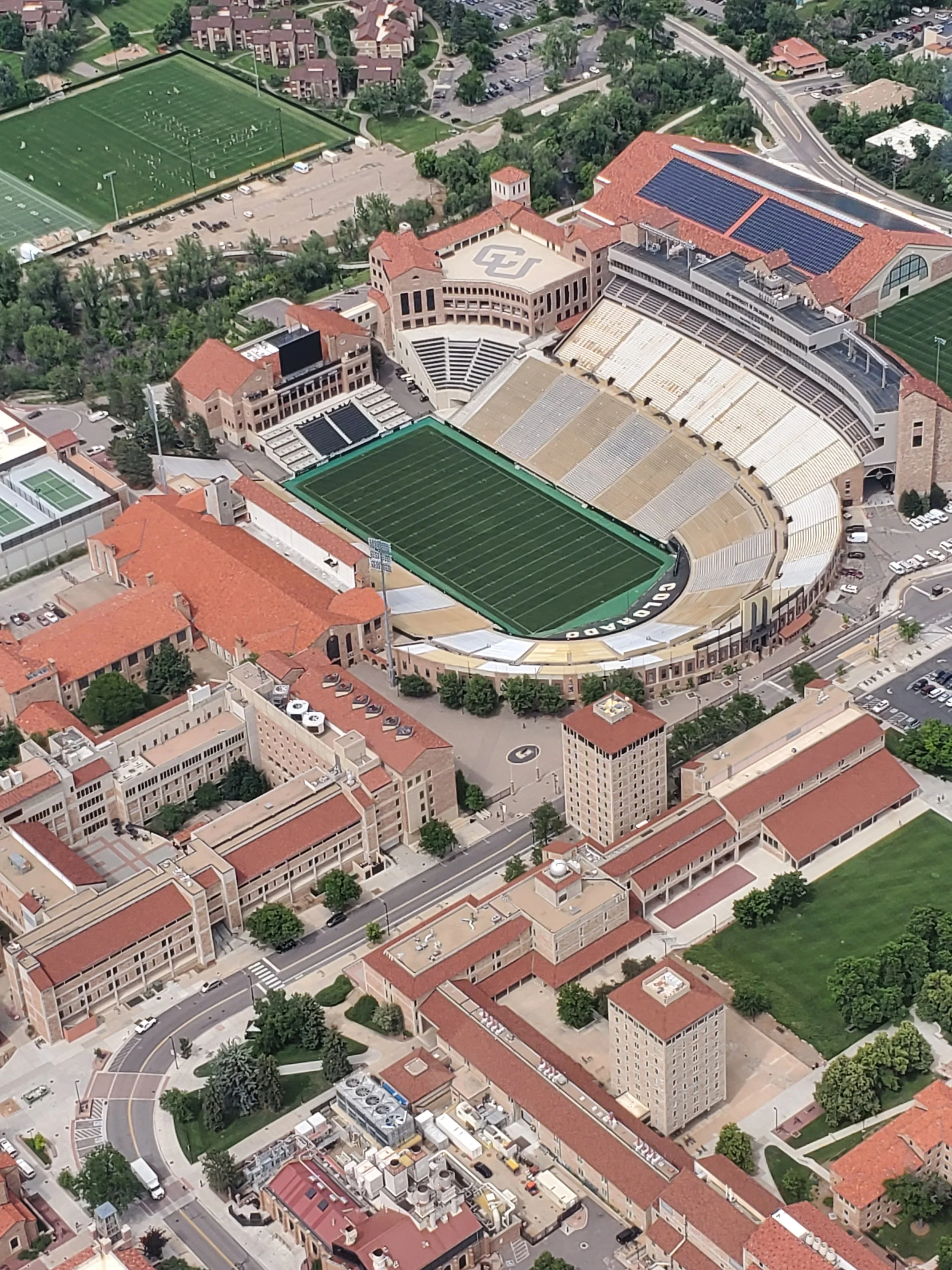 Aerial view of a university campus featuring a football stadium with a green field, surrounding buildings with red-tiled roofs, parking lots, and nearby sports fields.