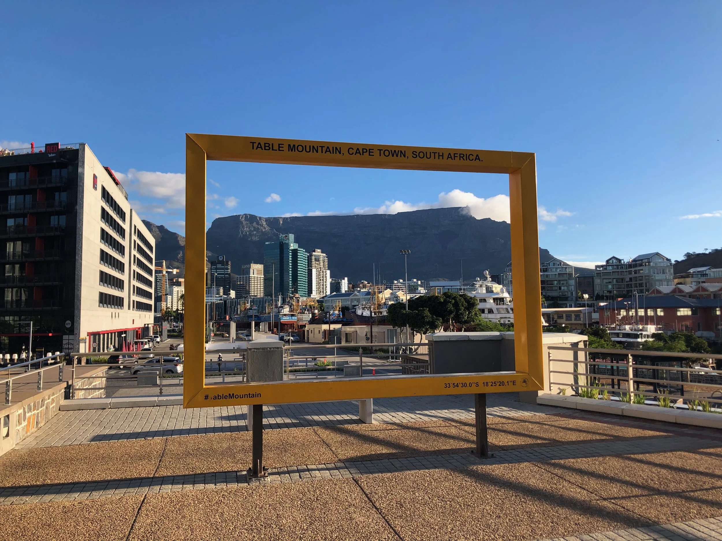 Urban scene featuring a yellow frame with the words 'Table Mountain, Cape Town, South Africa' written on top, city buildings, and a mountain backdrop on a clear day.