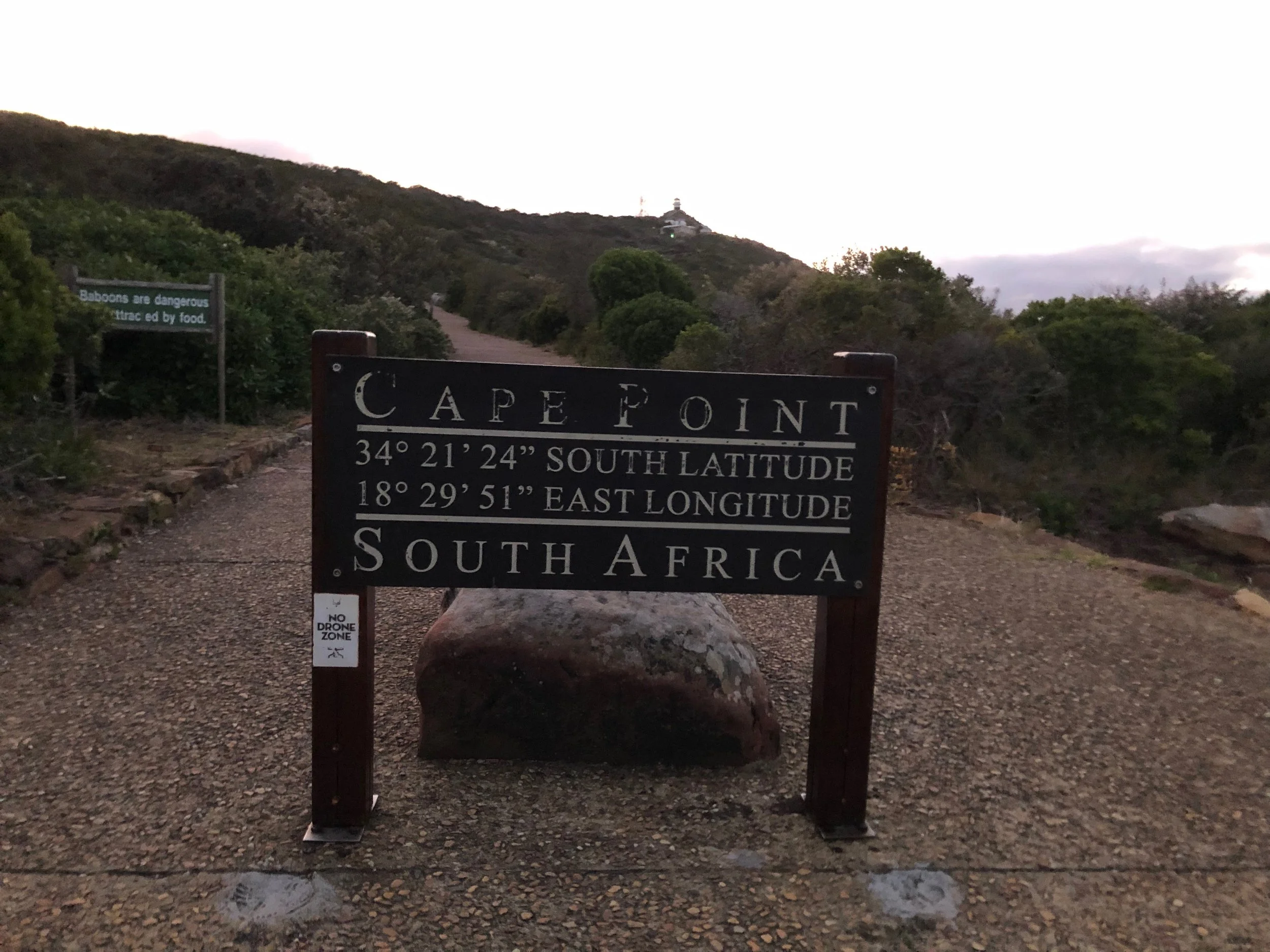 A sign at Cape Point with latitude and longitude coordinates, surrounded by greenery and a dirt path leading uphill, with a lighthouse visible in the distance against a cloudy sky.